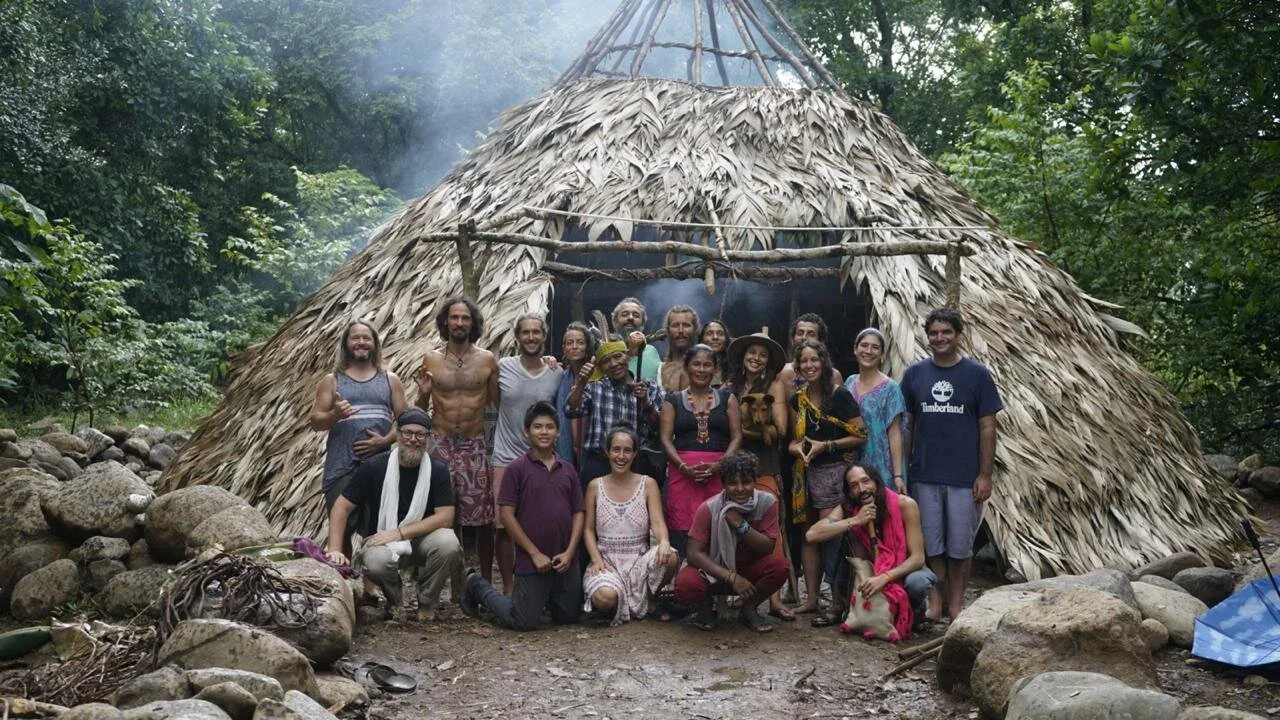 Group of people standing in front of a traditional thatched-roof hut in a jungle setting.