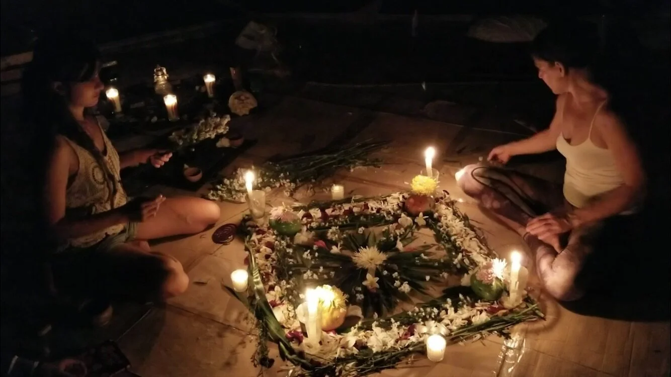 Two young women sit cross-legged on the floor in a dimly lit room, facing a large floral arrangement with candles. The scene suggests a memorial or spiritual ceremony with flowers and candles.