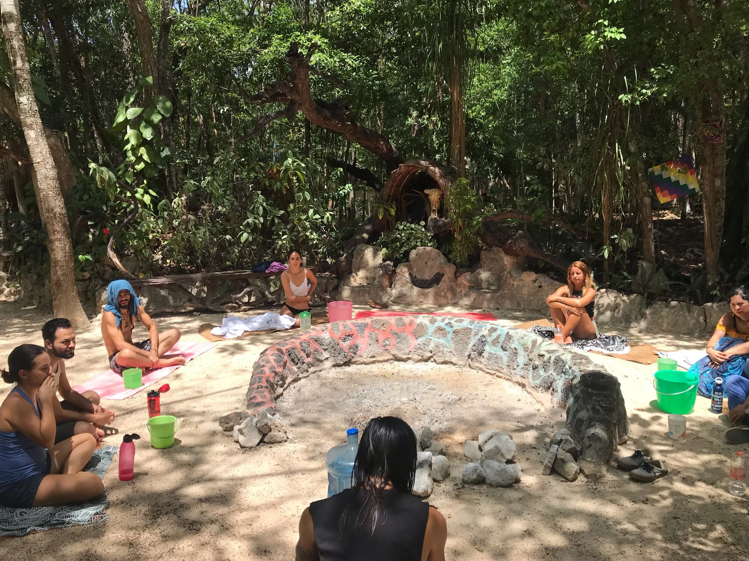 A group of people sitting cross-legged on the ground in a circle outdoors surrounded by trees, with a fire pit in the center, and various colorful buckets and bottles nearby.