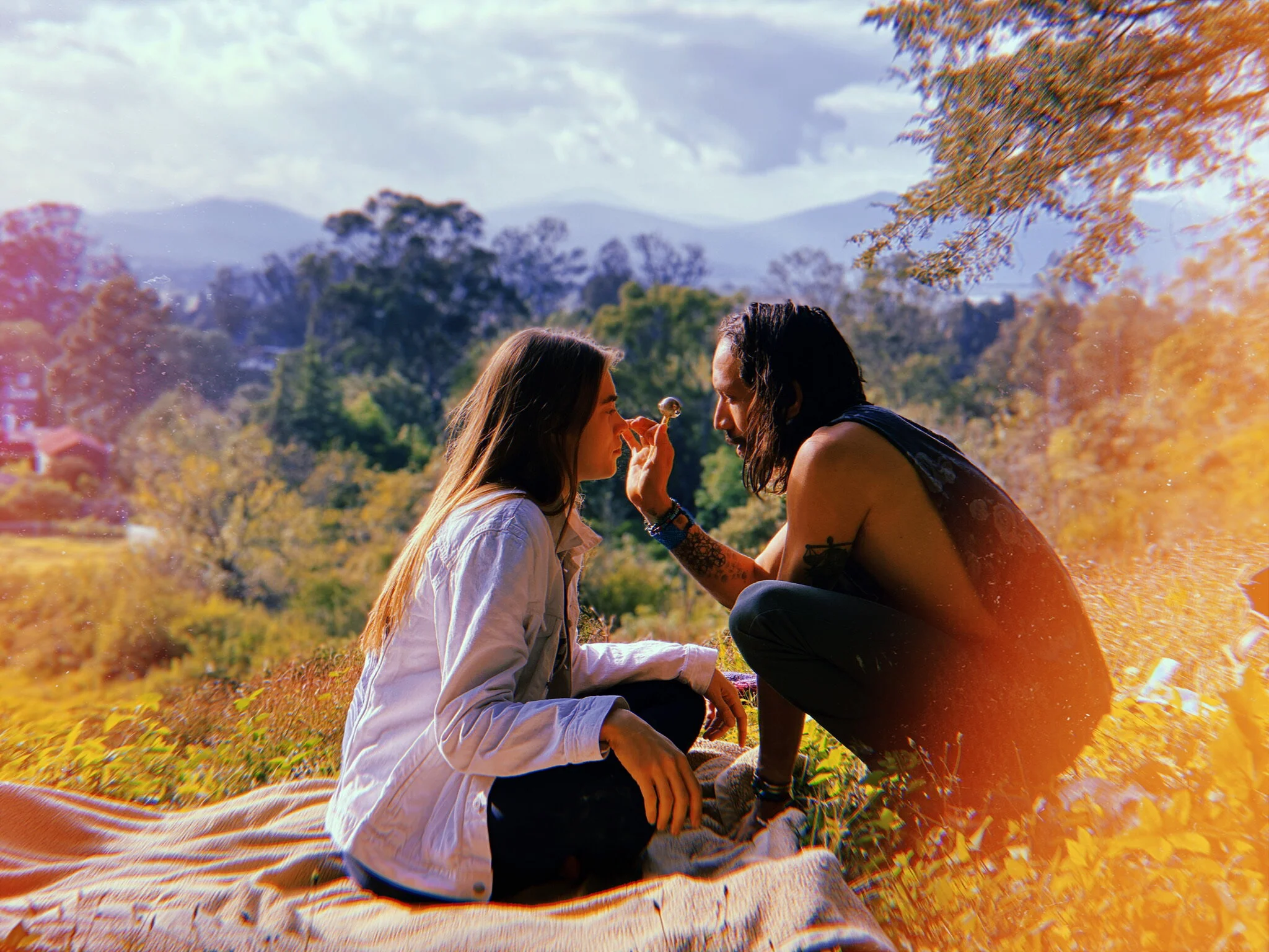 Two people, a young woman and a young man, sit on the grass with mountains and trees in the background. preparing for a bufo ceremony. breathwork first, connecting with the medicine. 