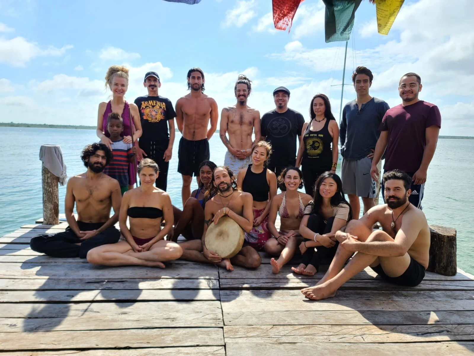 A diverse group of 17 people, including adults and children, smiling and sitting or standing on a wooden dock by a body of water on a sunny day. Some are wearing swimsuits, casual clothing, or yoga attire, with colorful flags hanging overhead.