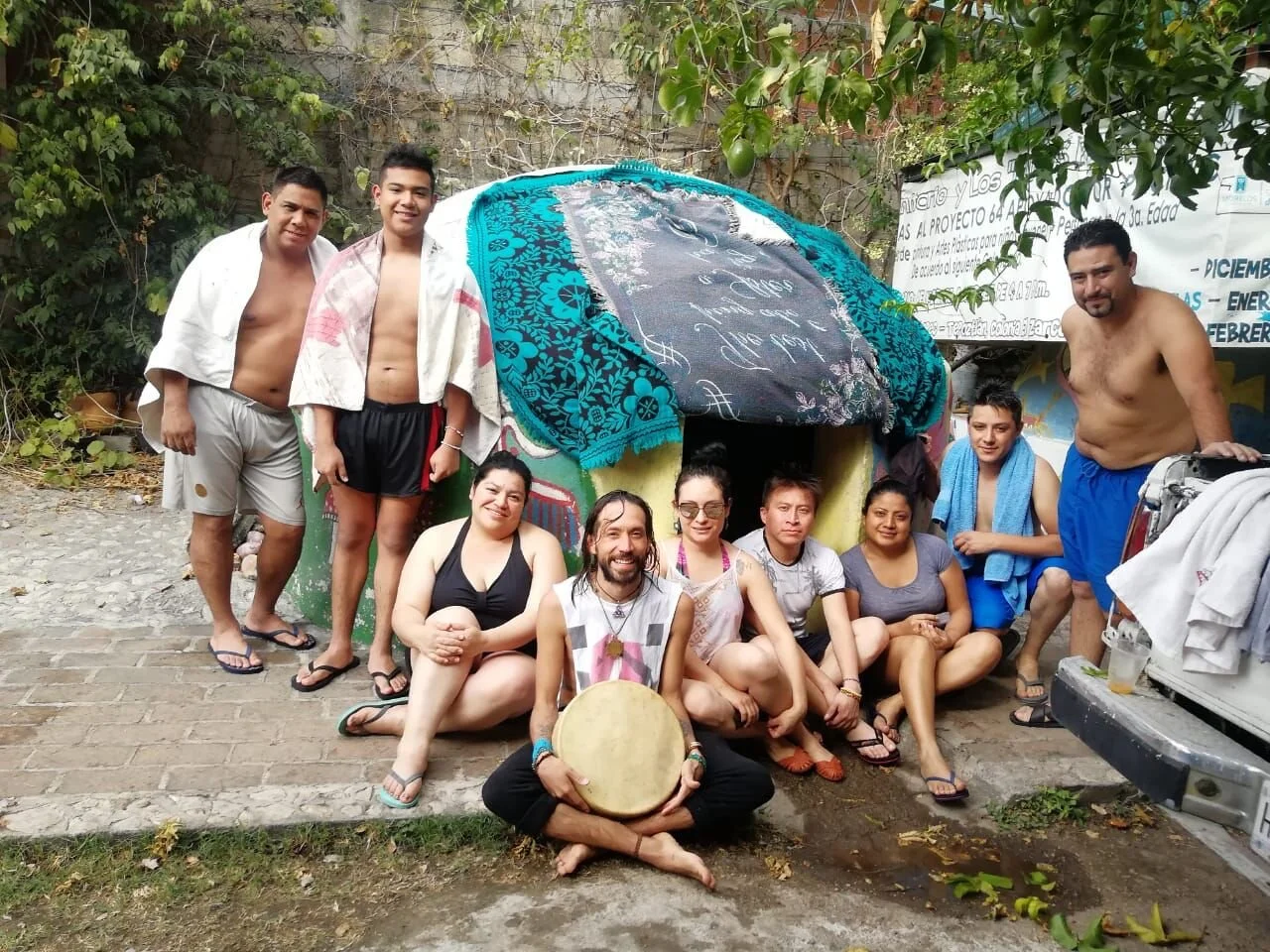 Group of people posing in front of colorful small house with a covered roof, some wrapped in towels, some in casual summer clothes, and one person holding a drum, outdoors surrounded by greenery.