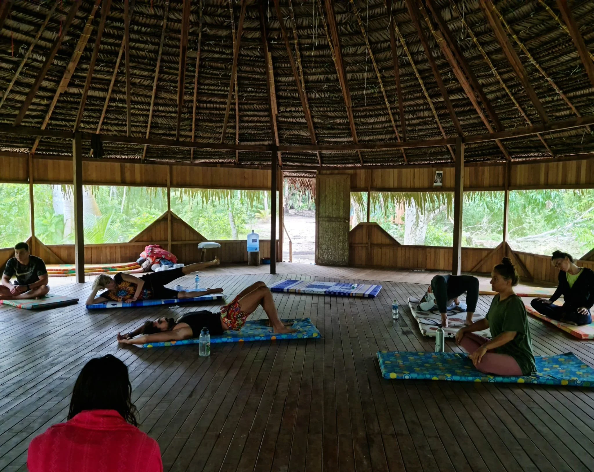 People practicing yoga or meditation on mats inside a wooden open-air structure with a thatched roof, surrounded by greenery.