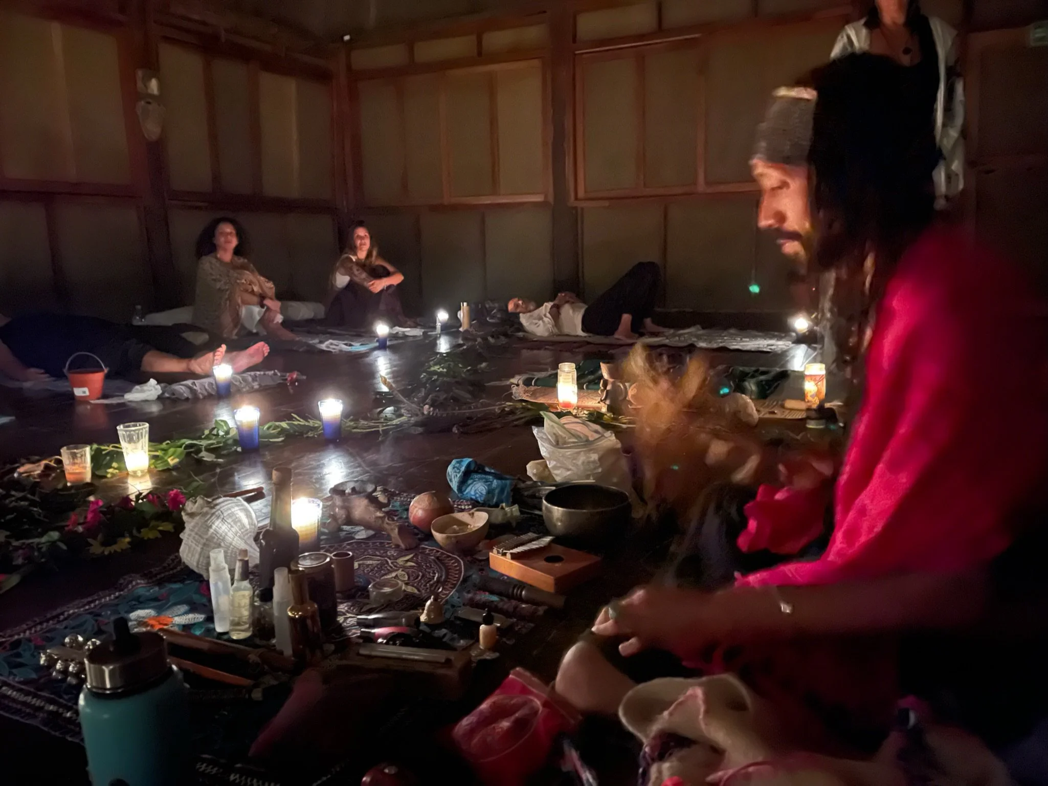 People sitting on the floor in a dimly lit room, surrounded by candles, flowers, and various objects, possibly participating in a meditation or spiritual gathering.