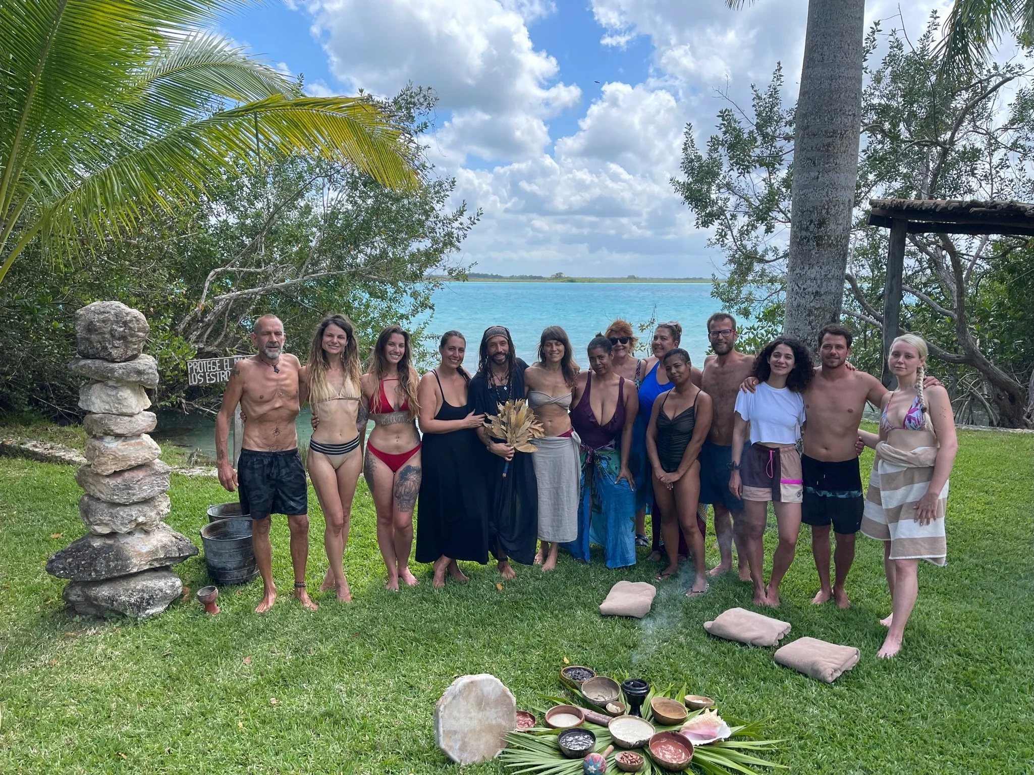 Group of people in swimsuits and casual clothing standing on grass near water, with trees and a cloudy sky in the background, participating in a ceremony or gathering.