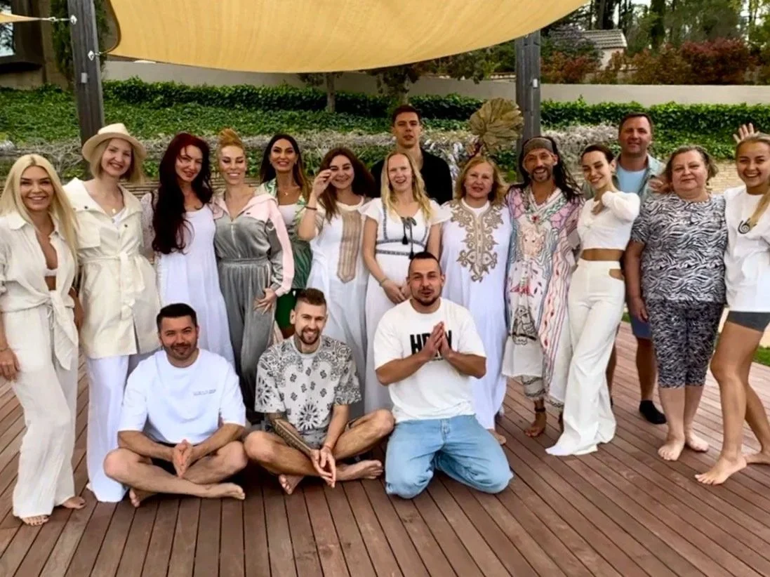 Group of 16 people posing outdoors on a wooden deck under a shade sail.