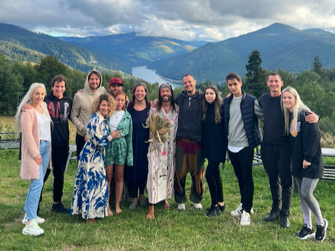 Group of thirteen people standing outdoors on grassy area with mountains and a lake in the background, some holding bouquets, smiling at the camera.