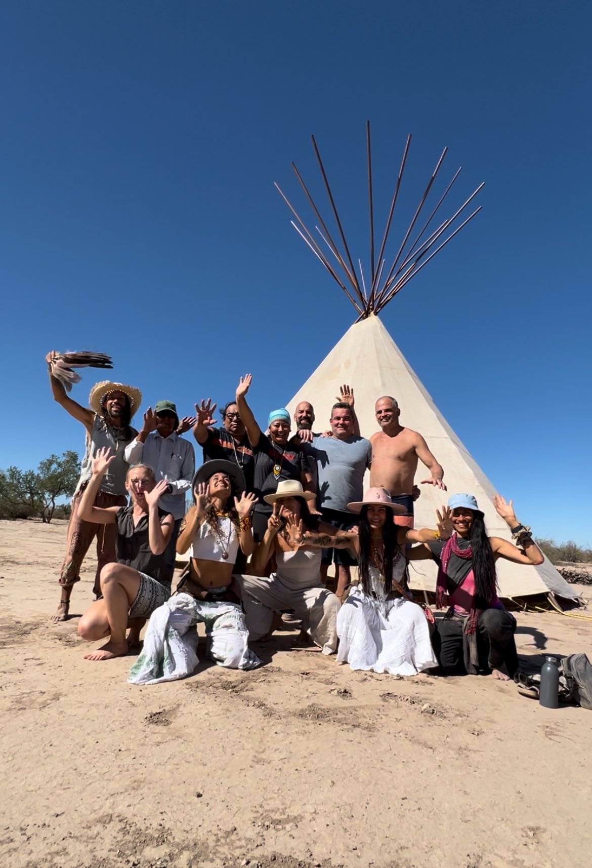Our diverse & happy group celebrating their journey  in Sonora Desert Bufo Alvarius Retreat in front of a tipi tent with a large teepee structure and a clear blue sky.
