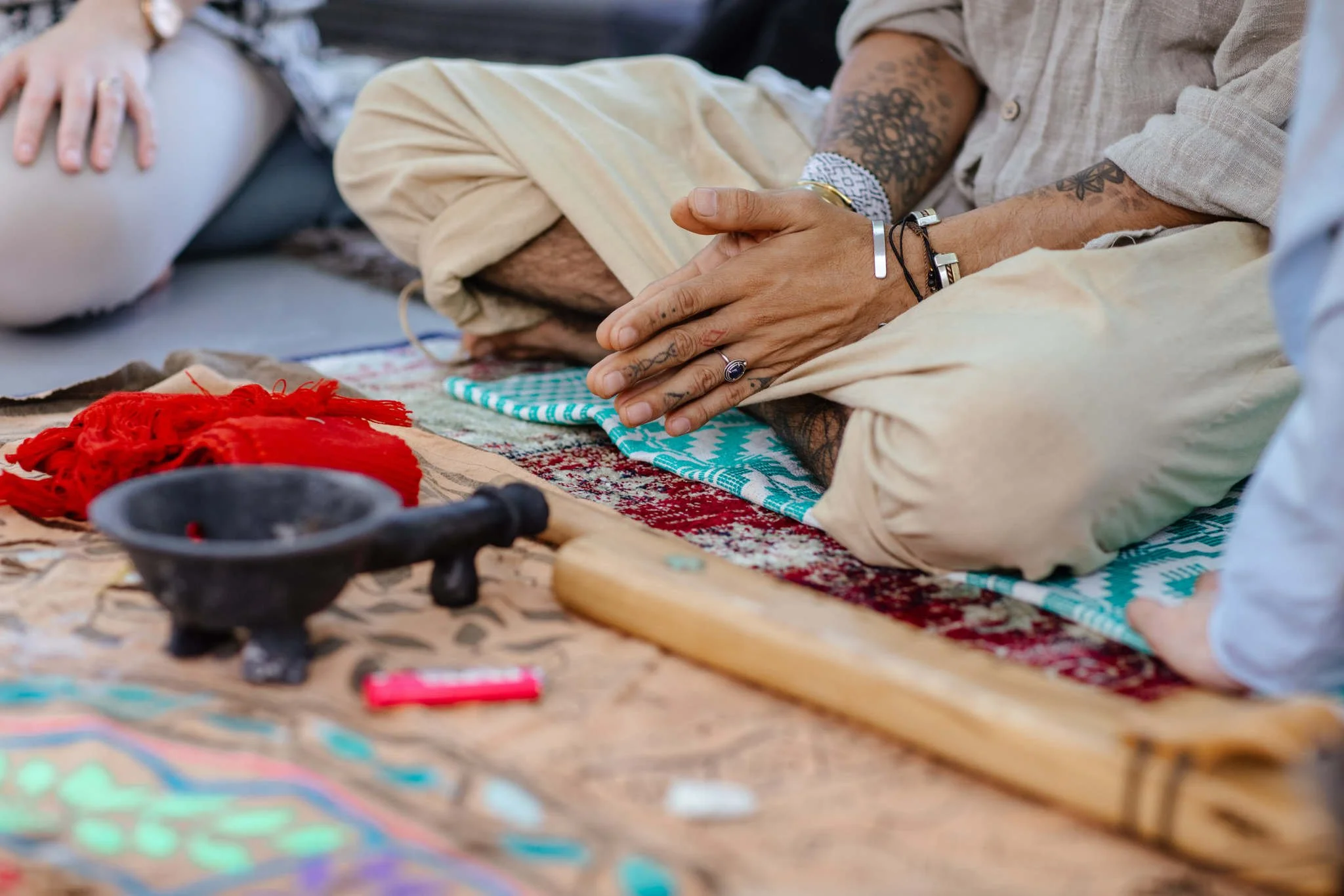 Close-up view of a person's hands with tattoos, wearing bracelets and rings, resting on their lap. The person is sitting cross-legged on woven fabric, with various objects and a red cloth visible in the foreground.