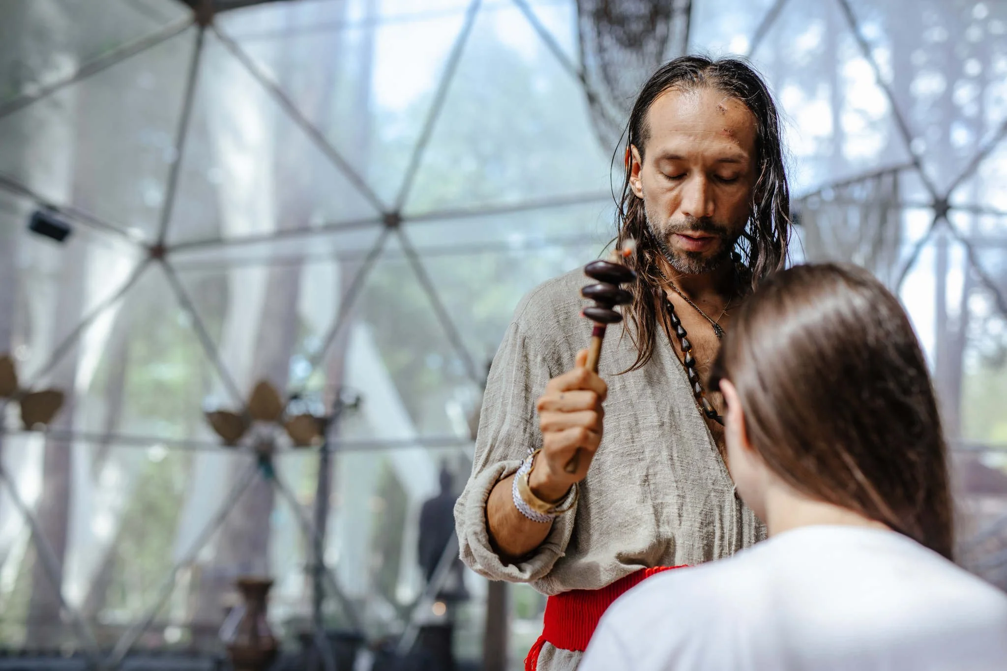 A woman with long dark hair and a beaded necklace performing a healing or spiritual ritual on a child, dressed in a white shirt with a red sash, inside a geodesic dome with a blurry background of trees.