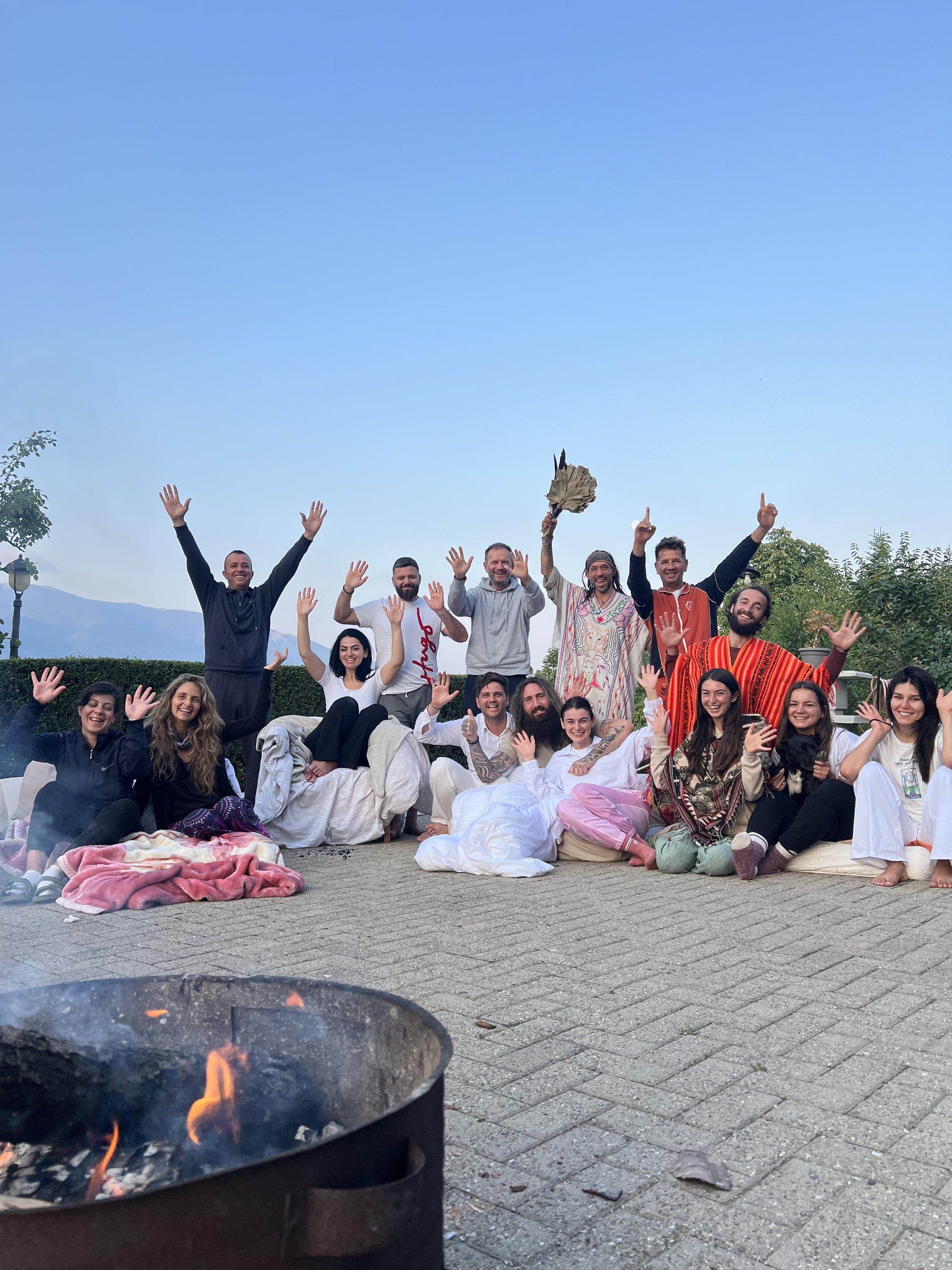 A happy group of people gathered, smiling after our peyote ceremony, with a fire pit and mountains in the background.