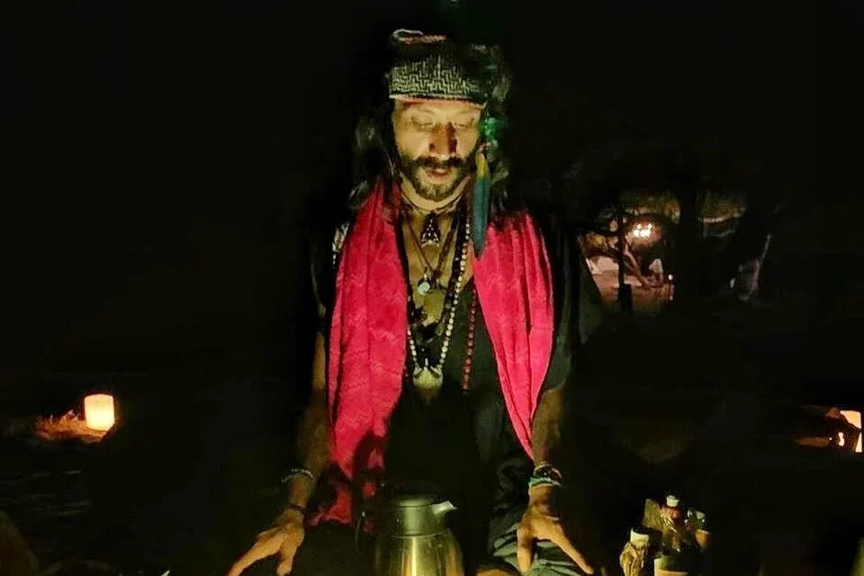 A man dressed as a mystical or pirate character, with long hair, a headband, and layered necklaces, standing in a dark outdoor setting with candles in the background.