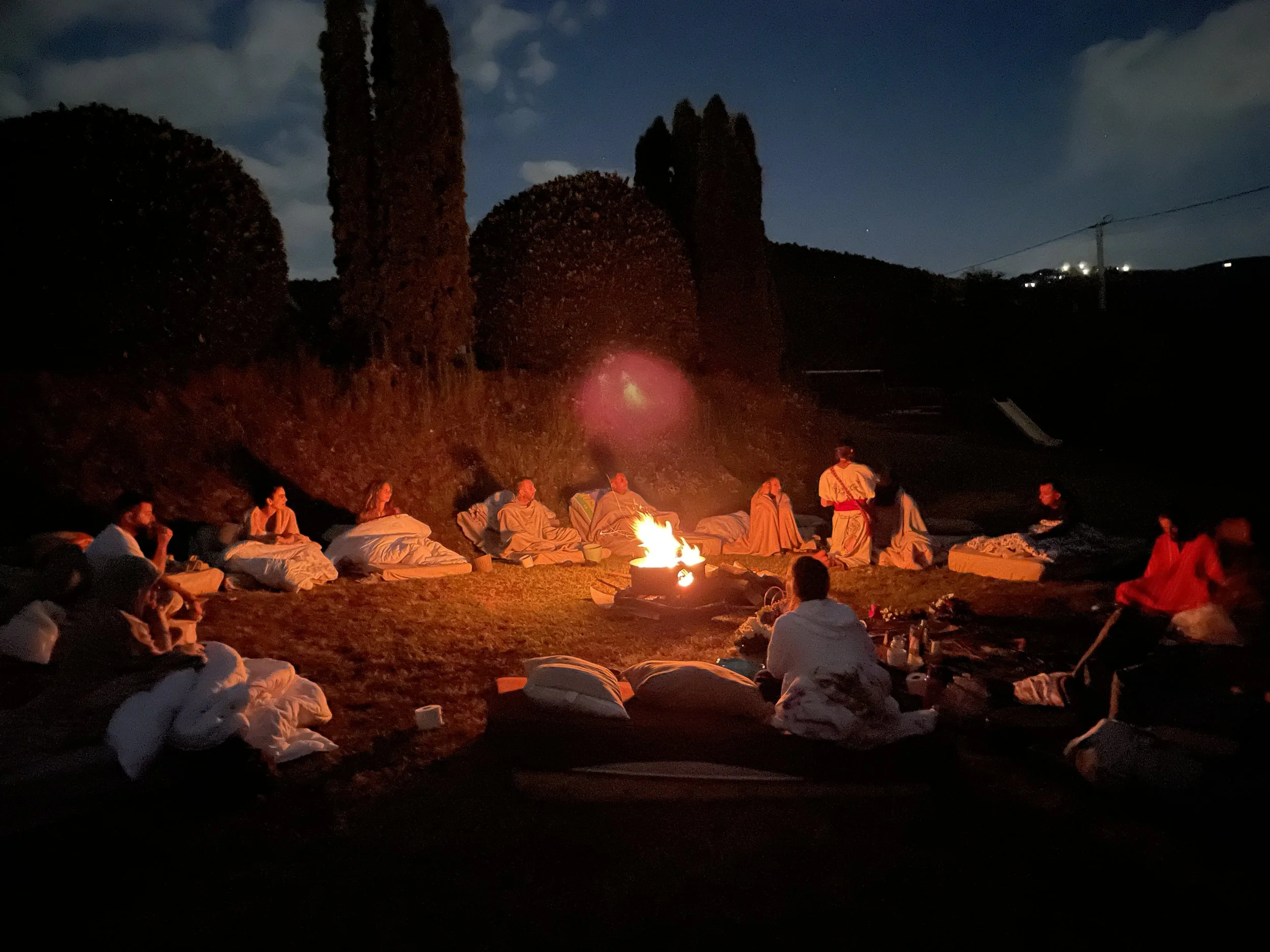 People sitting around a campfire at night for peyote ceremony in mexico with tall trees and a starry sky in the surrounding us.