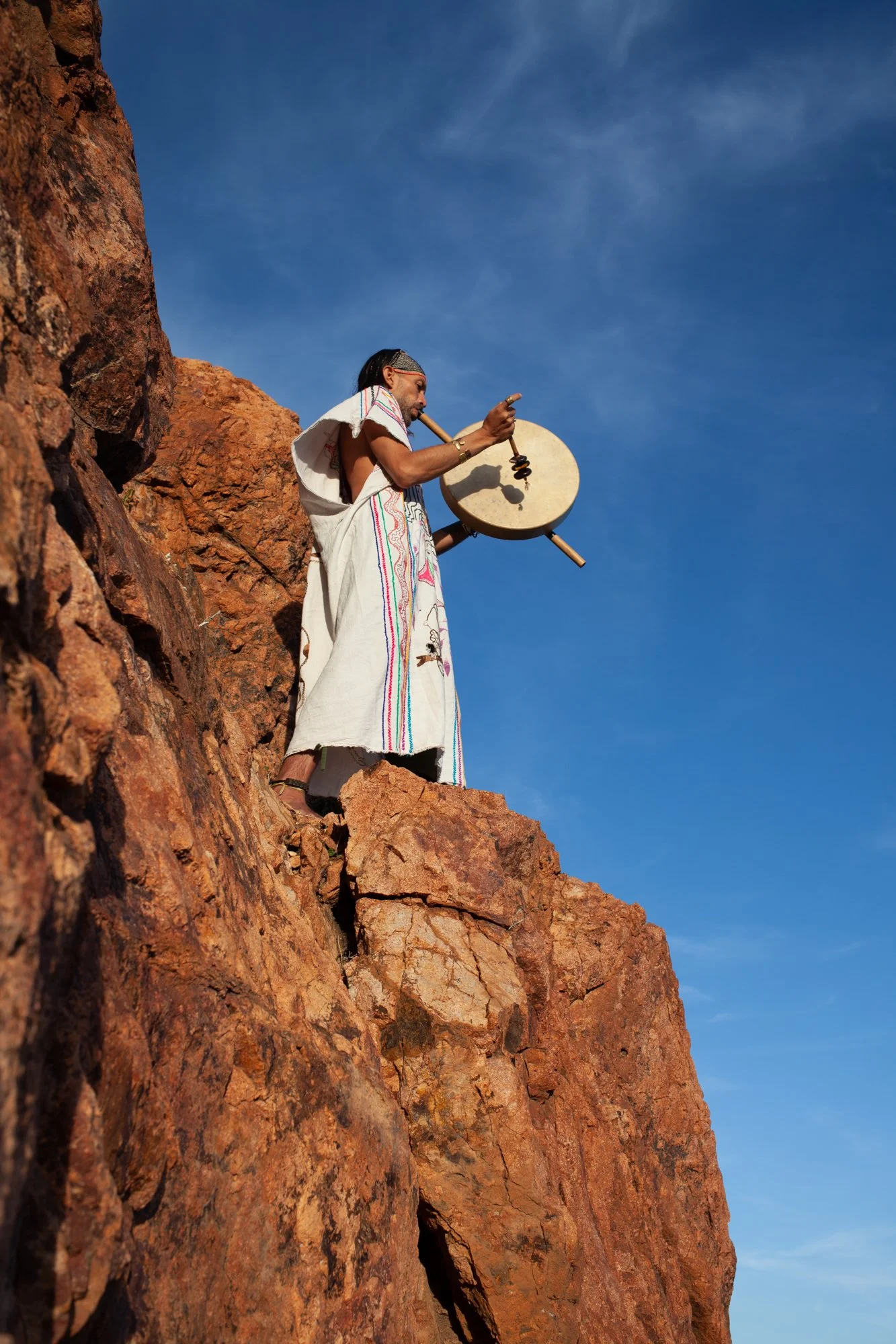 A person standing on red rocky terrain, playing a traditional drum, wearing traditional clothing with colorful embroidery and a headband, against a blue sky with wispy clouds.