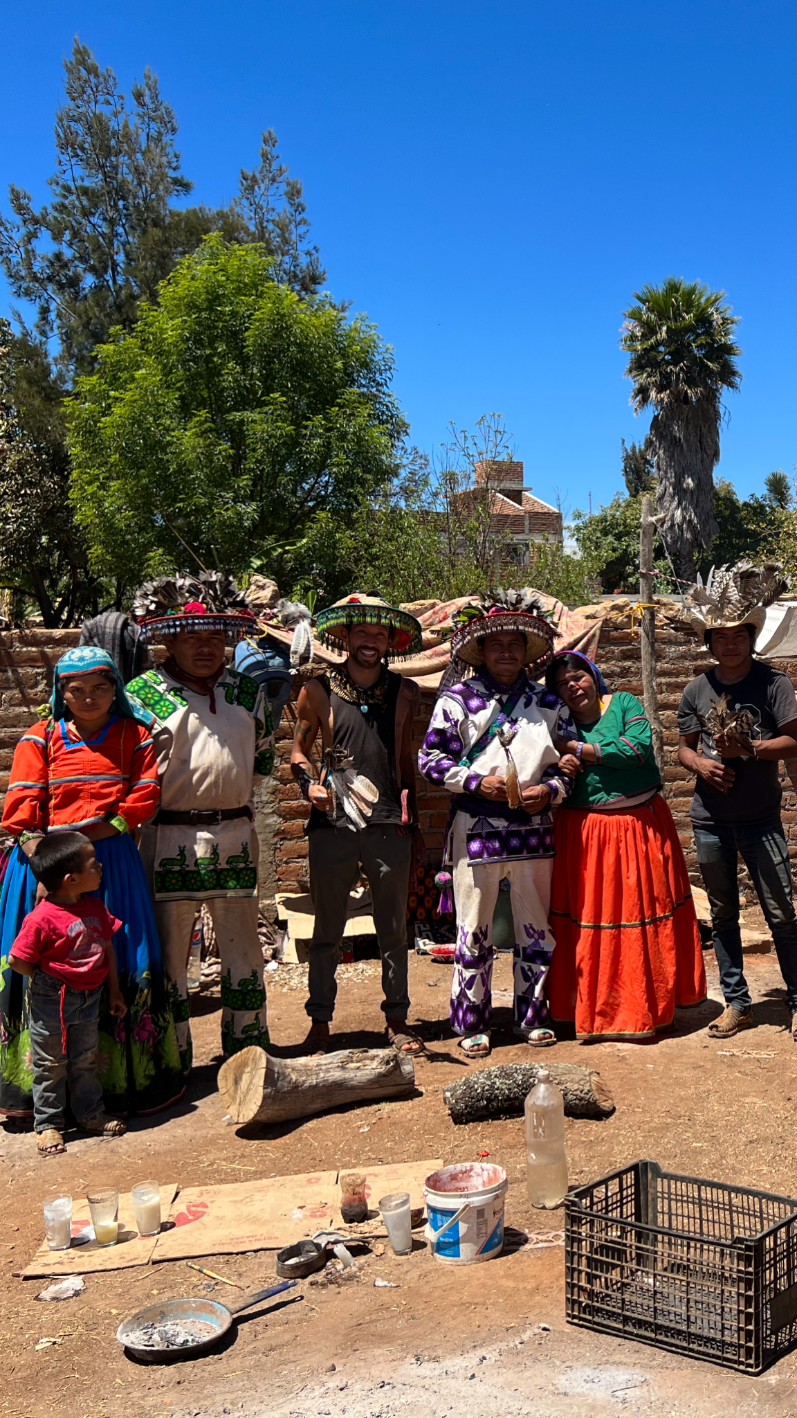 A group of people dressed in traditional Mexican attire, standing outdoors in front of a brick wall, with trees and a house in the background. The scene appears to be part of a cultural event or celebration.
