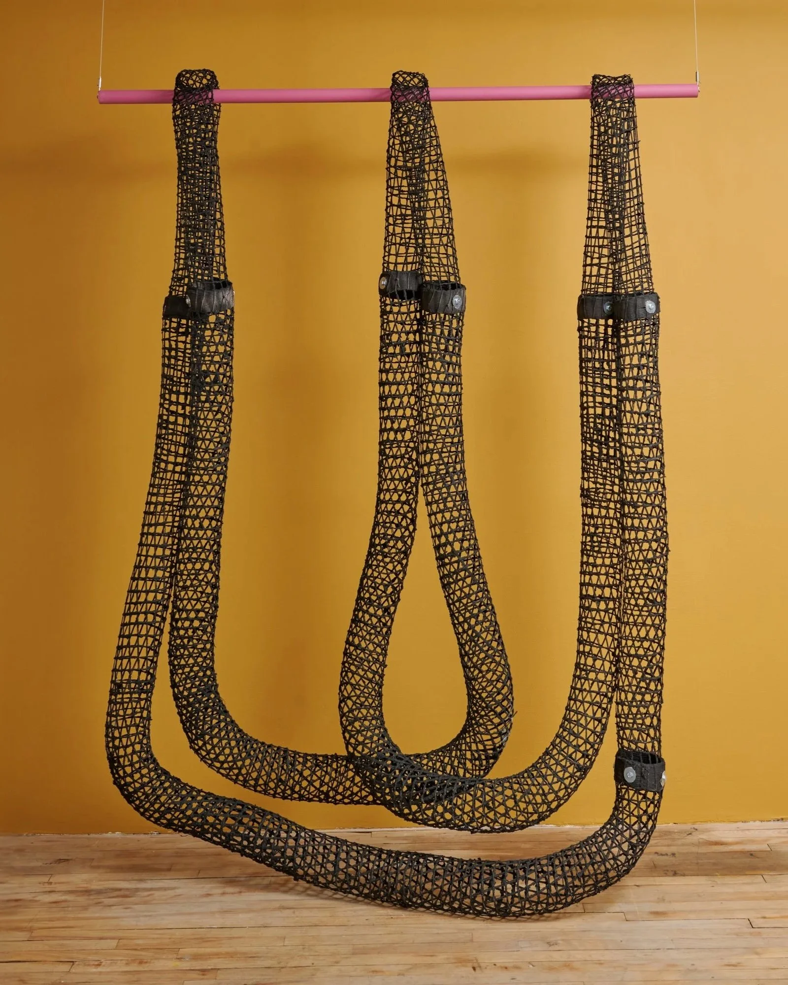 Three black mesh belts hanging from a pink rod against a yellow wall.