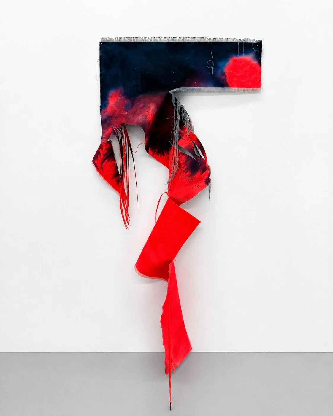 An art piece featuring a large, abstract letter 'F' with a red, black, and dark blue color palette, made of mixed media materials that include fabric and paint, displayed against a white wall. Vaughn Davis Jr.