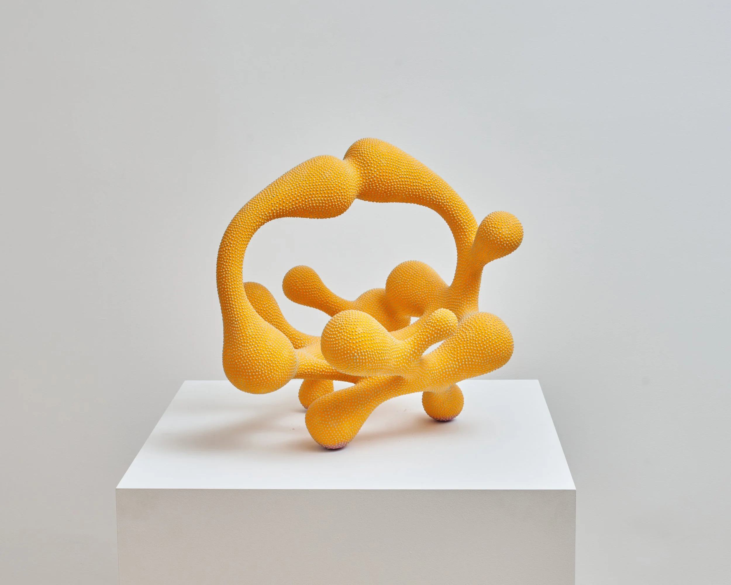 A modern sculpture resembling abstract human hands in bright yellow with orange accents, set on a white pedestal against a gray background. Joël Brodovsky-Adams.