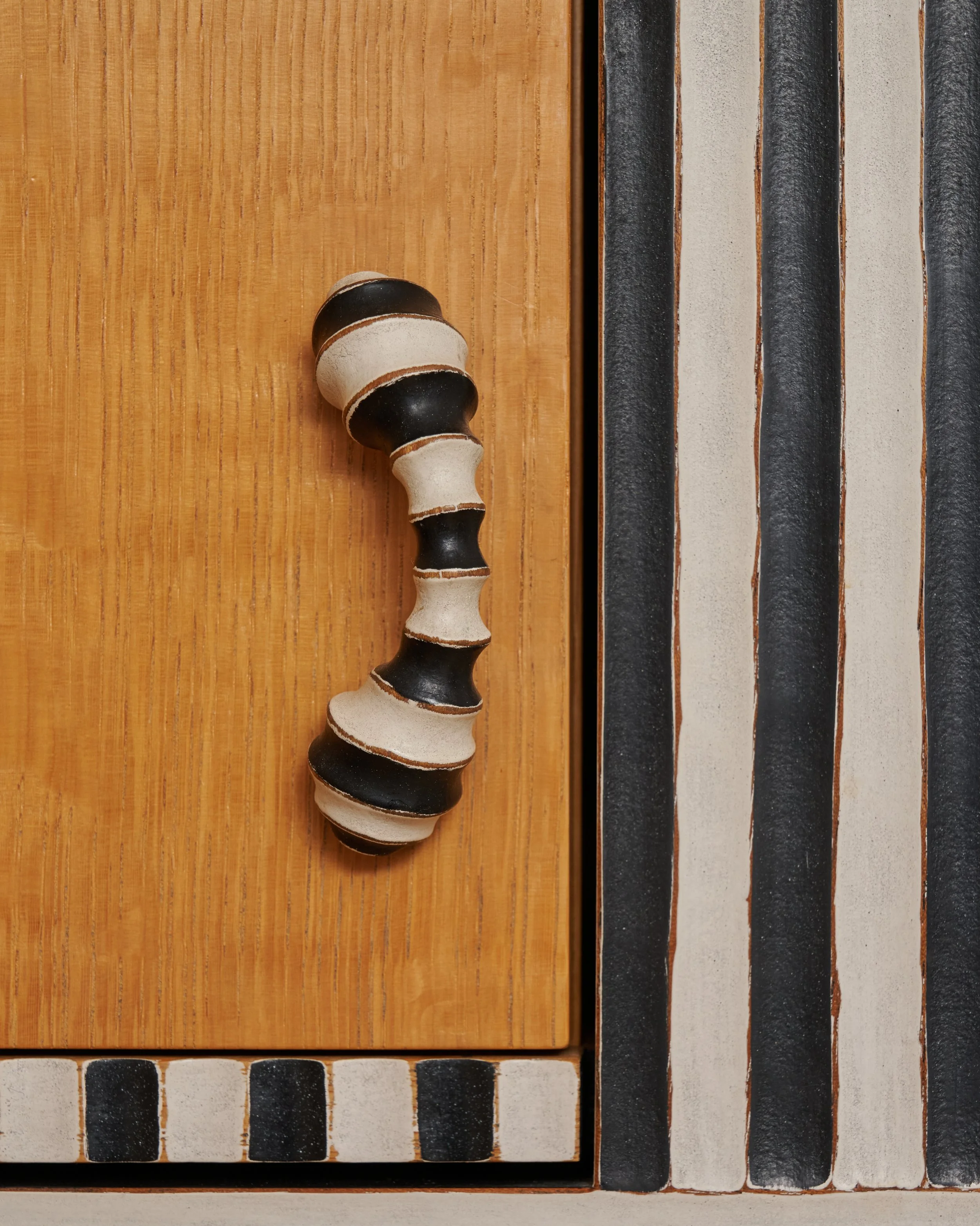 A wooden tabletop with a black, white, and gray striped ceramic handle on the left side, and a wooden border on the edge, with a part of a tiled surface with black and white striped tiles visible at the bottom. Tom Loeser.