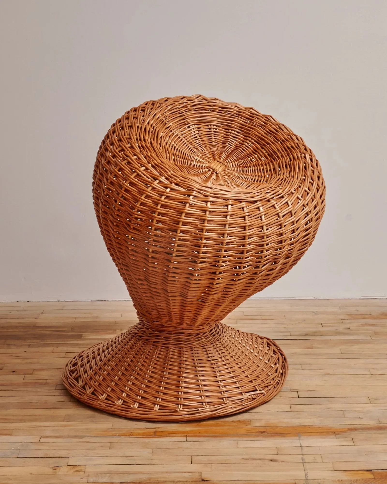 A large, abstract wicker sculpture resembling a human head, placed on a wooden floor against a plain wall.
