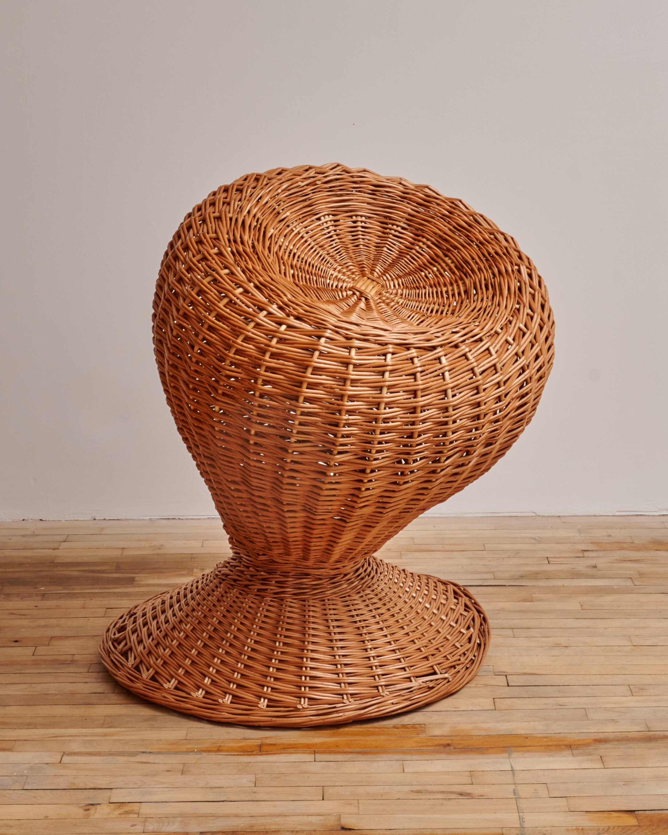 A large, woven wicker chair with a rounded, bowl-shaped seat and a wide, flat base, placed on a wooden floor against a plain white wall.