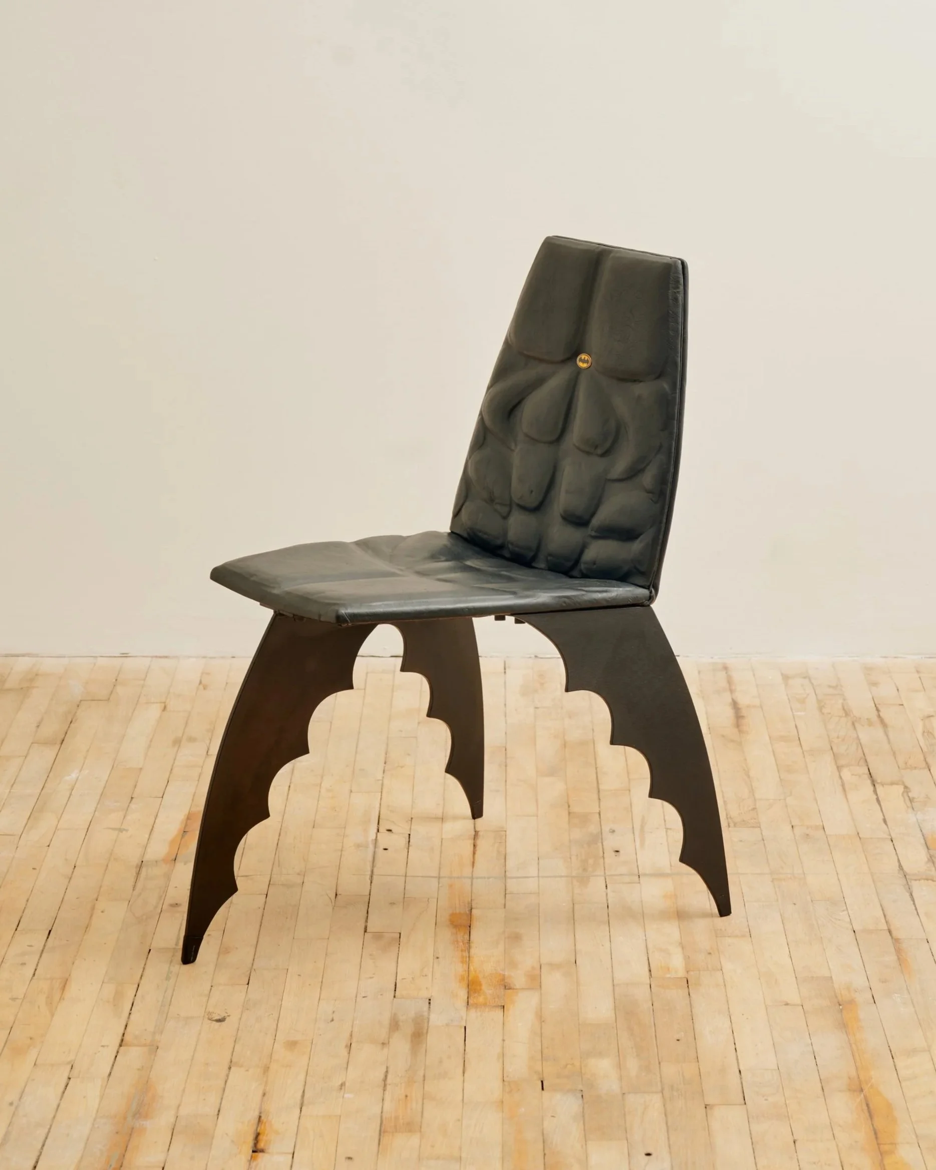 A black leather chair with a tall back and carved wooden legs, placed on a light wooden floor against a plain white wall.