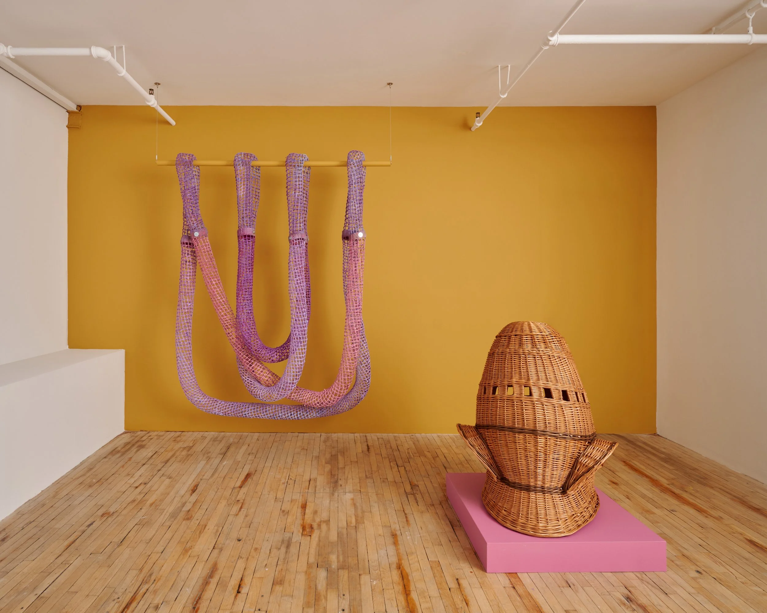 Art installation featuring a large, colorful rope sculpture hanging from the ceiling and a woven wicker chair on a pink platform against a yellow wall in a gallery space.