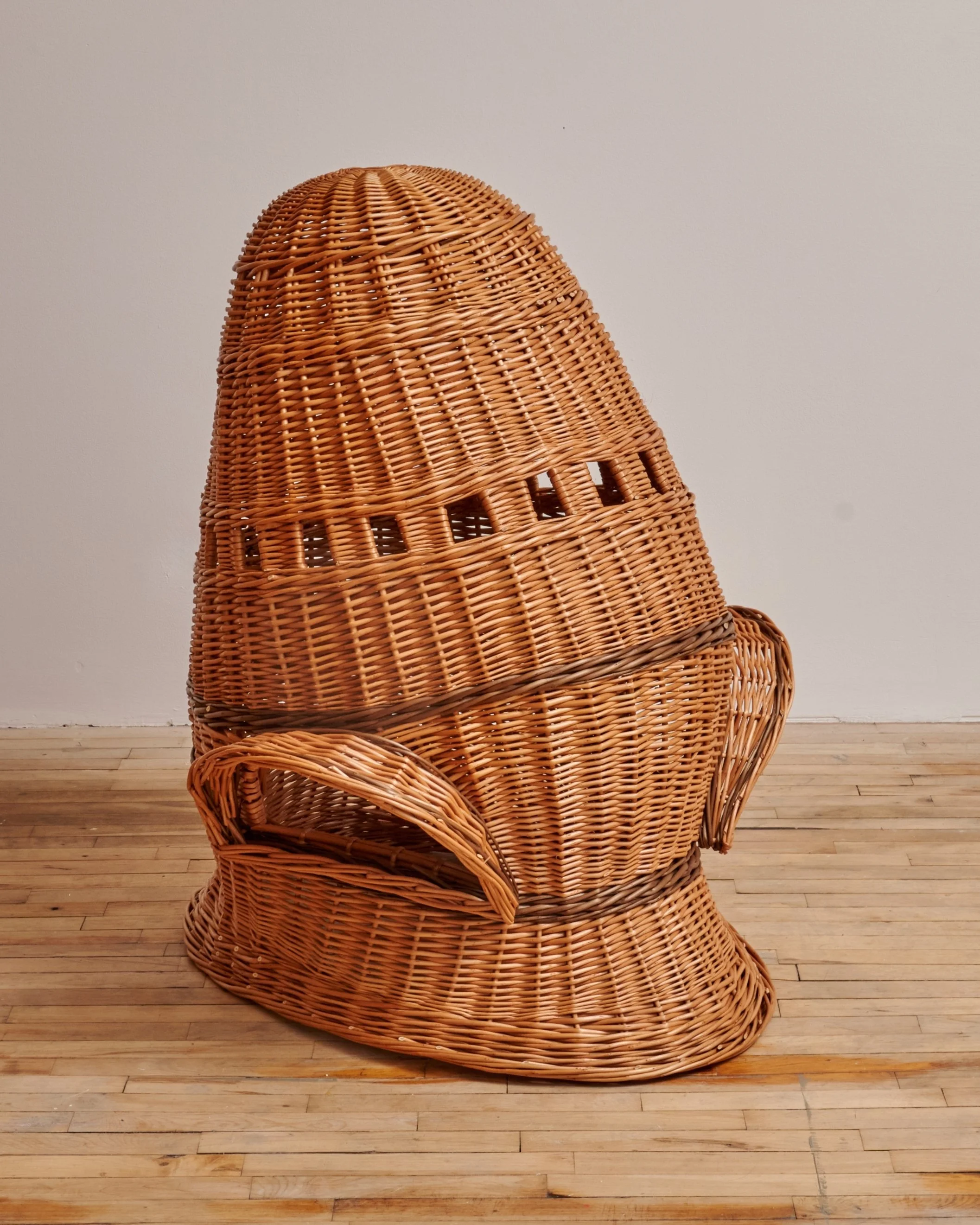 A sculptural art piece made of woven wicker in the shape of a rocket ship, set against a plain white wall and placed on a wooden floor.