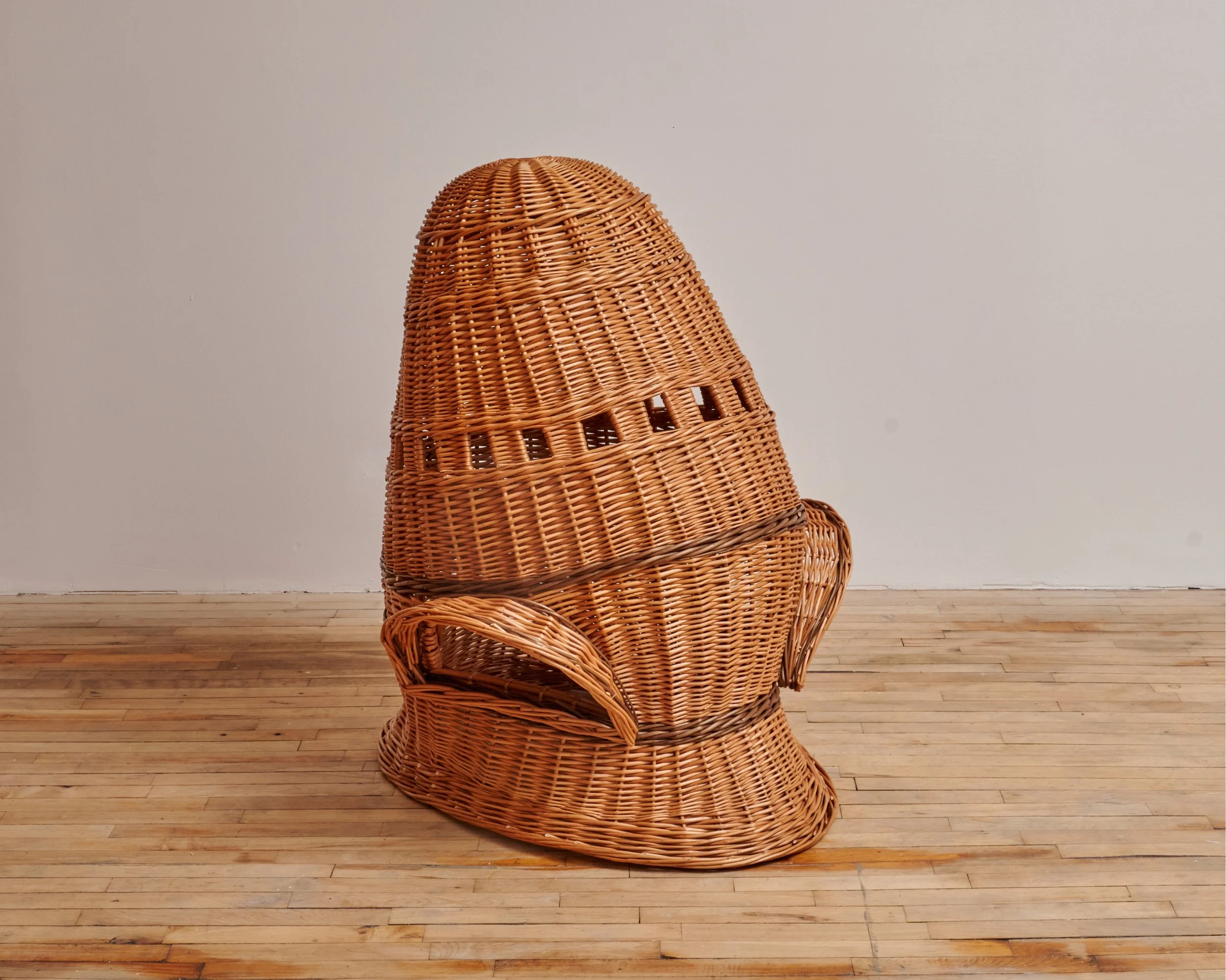 A chair made entirely of woven wicker in the shape of a rocket placed on a wooden floor against a plain wall.