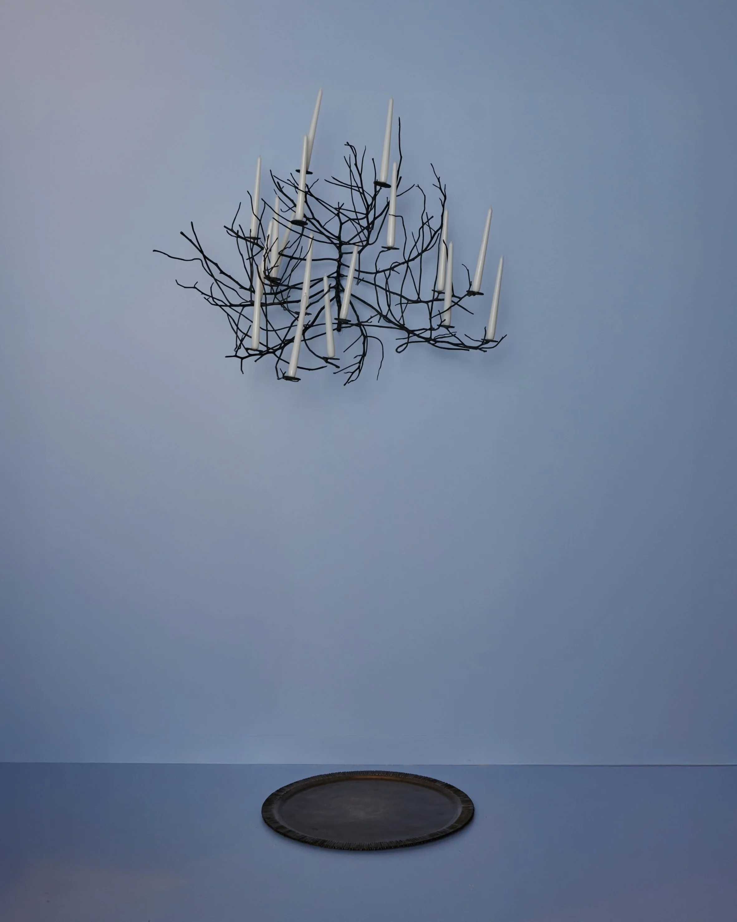 A metal wall sculpture resembling a chandelier with candle-like extensions, hanging above a dark circular table or tray on a blue wall.