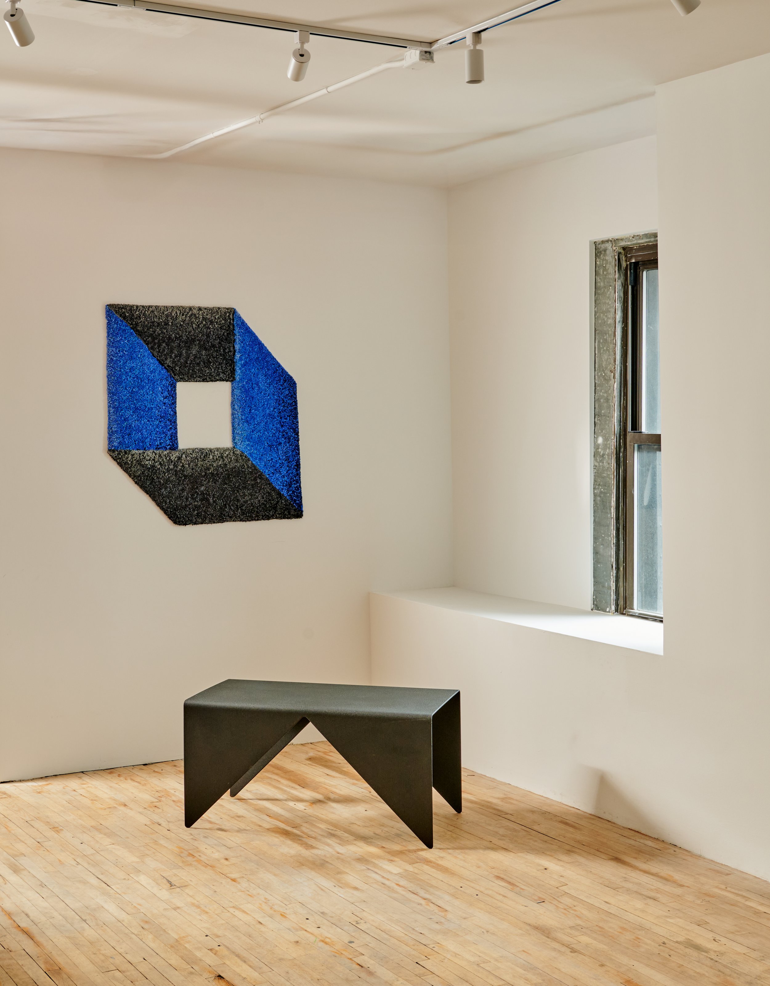 Minimalist art installation in a gallery featuring a wall-mounted geometric piece with black and blue colors and a black angular sculptural piece on the wooden floor near a window.