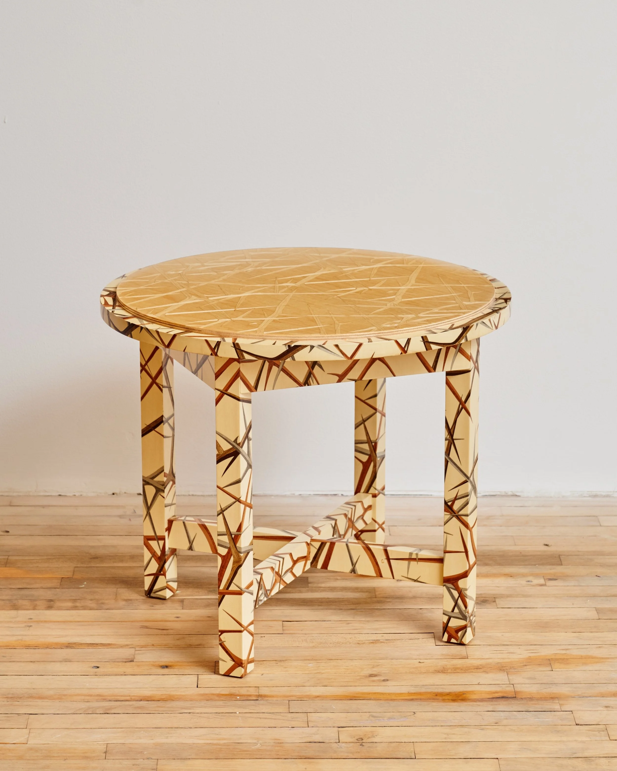 A round wooden table with a unique geometric pattern on its surface and legs, placed against a white wall and on a wooden floor.