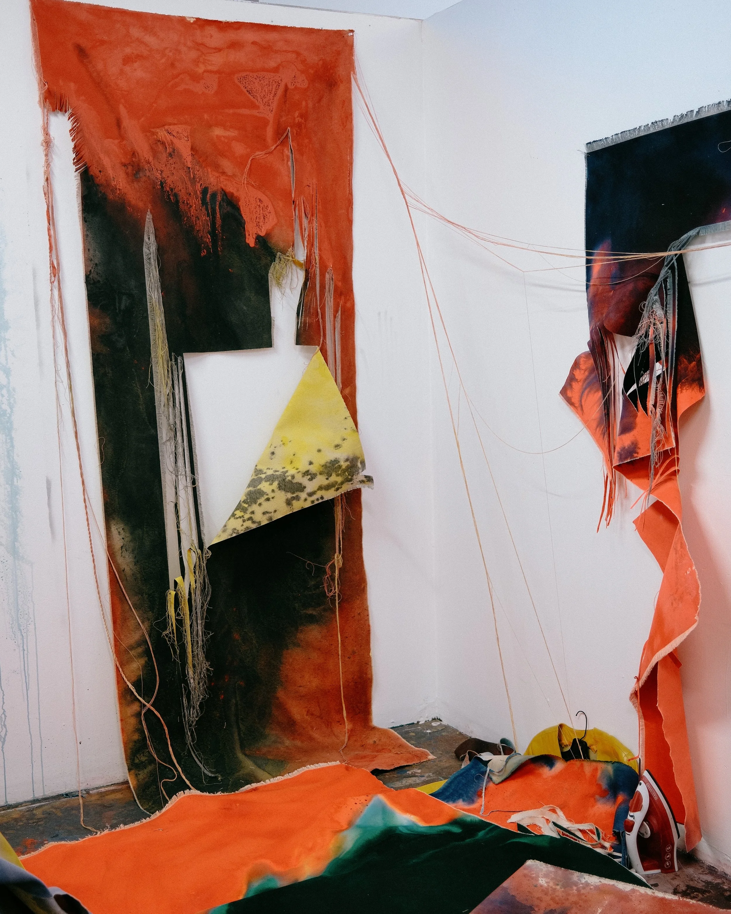 An art installation in a gallery featuring large, abstract fabric pieces with fiery orange and black colors, hanging from the ceiling and draping onto the floor, with visible orange strings connecting the fabrics to the ceiling. Vaughn Davis Jr.