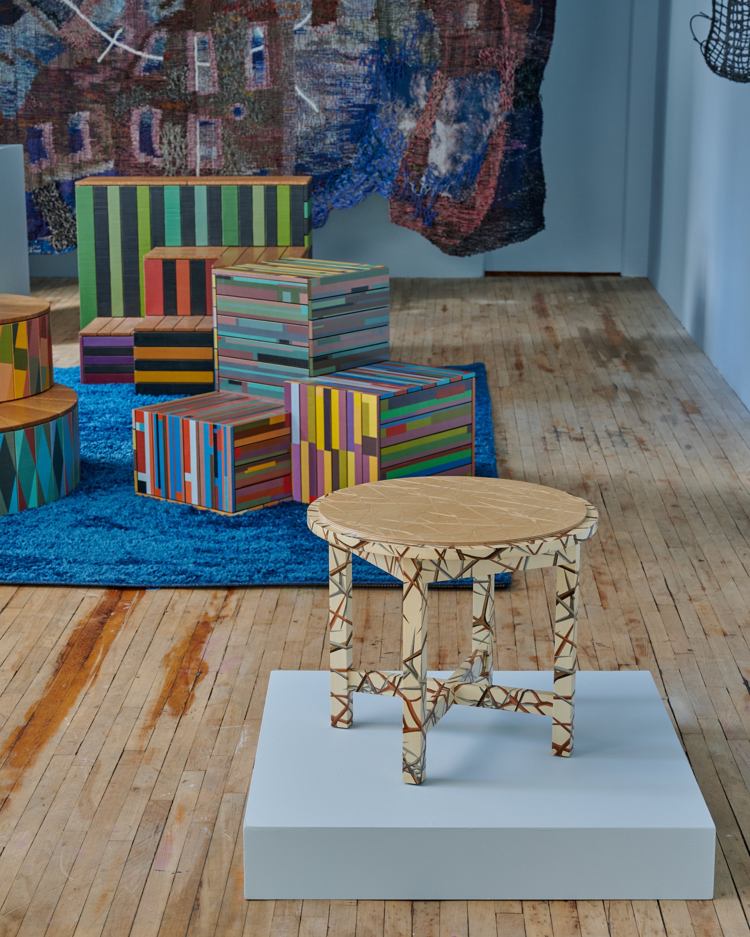 A small mosaic-style wooden table on a white pedestal in an art gallery, with colorful boxes and a blue rug in the background.
