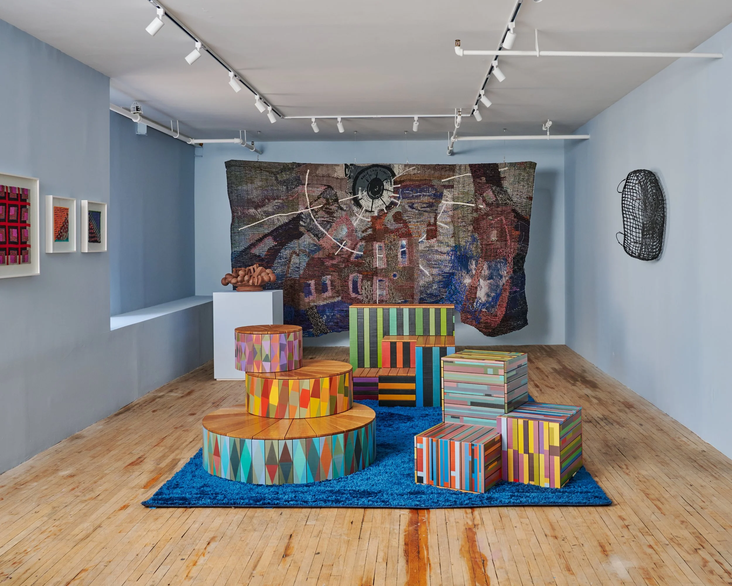 An art gallery with a large, abstract textile artwork hanging on a light blue wall. There are smaller framed artworks on the left wall, a black wire basket on the right wall, and a white pedestal with a clay sculpture on top. In the center, multi-colored, geometric-patterned furniture pieces are arranged on a blue rug, set against a wooden floor.