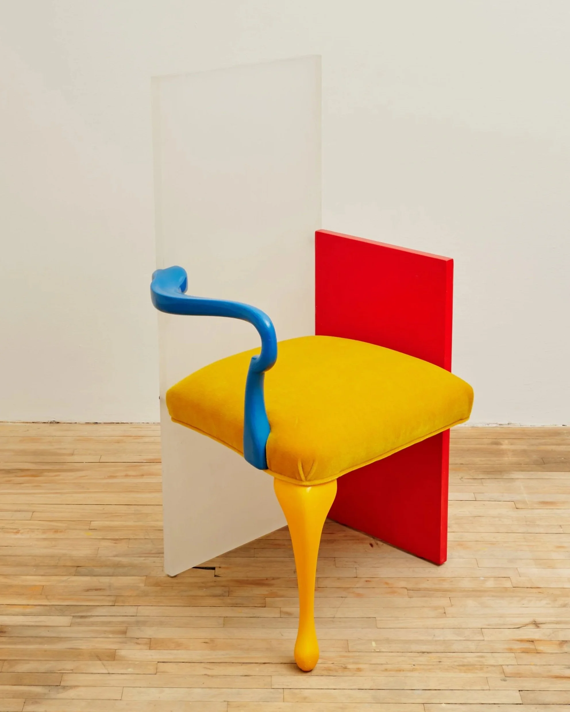 An abstract chair with a yellow cushioned seat, a red backrest, blue armrest, and a yellow leg, positioned against a white wall on a light wooden floor.