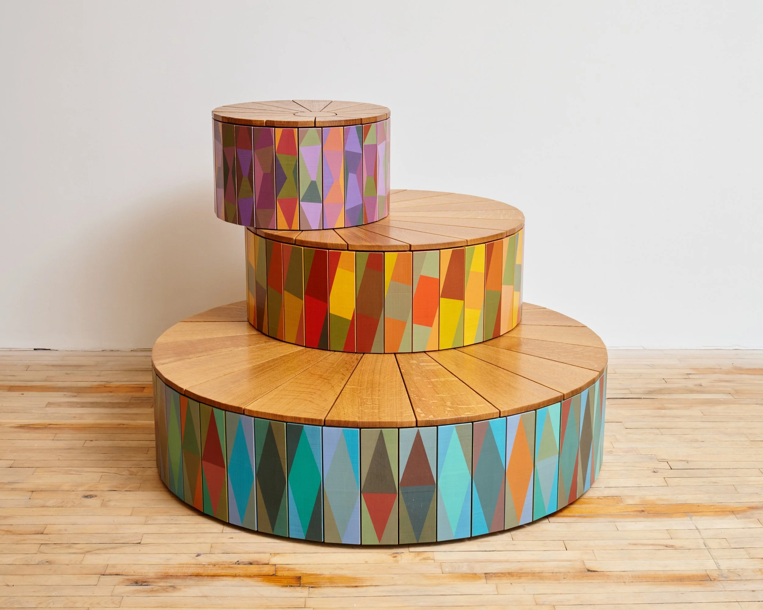 Three-tiered wooden circular platforms with colorful tile patterns stacked in a stepped arrangement on a wooden floor against a white wall.