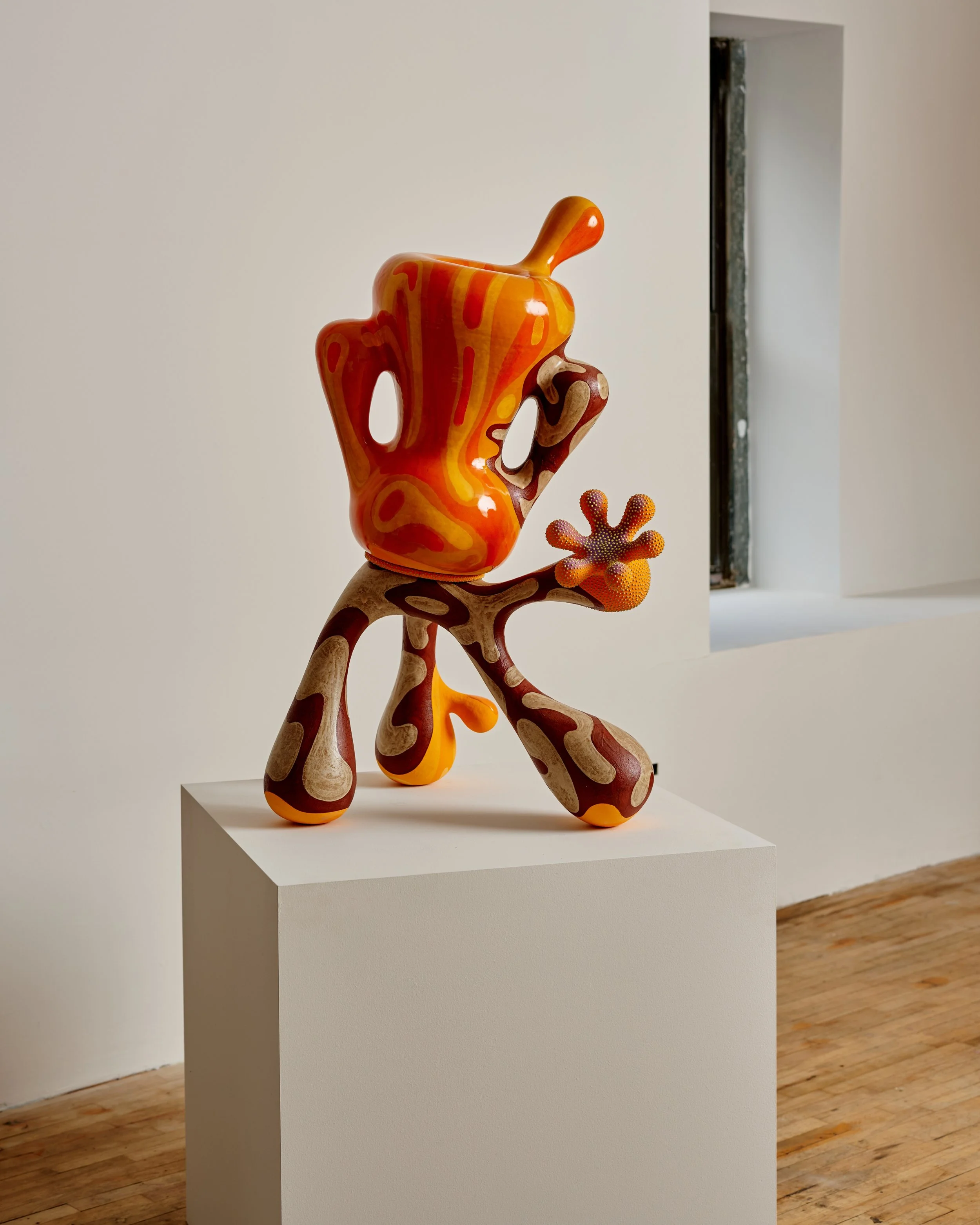 Colorful ceramic sculpture resembling a stylized animal or creature with elongated limbs, rounded body, and decorative patterns, displayed on a white pedestal in an art gallery with white walls and wooden floor. Joël Brodovsky-Adams.