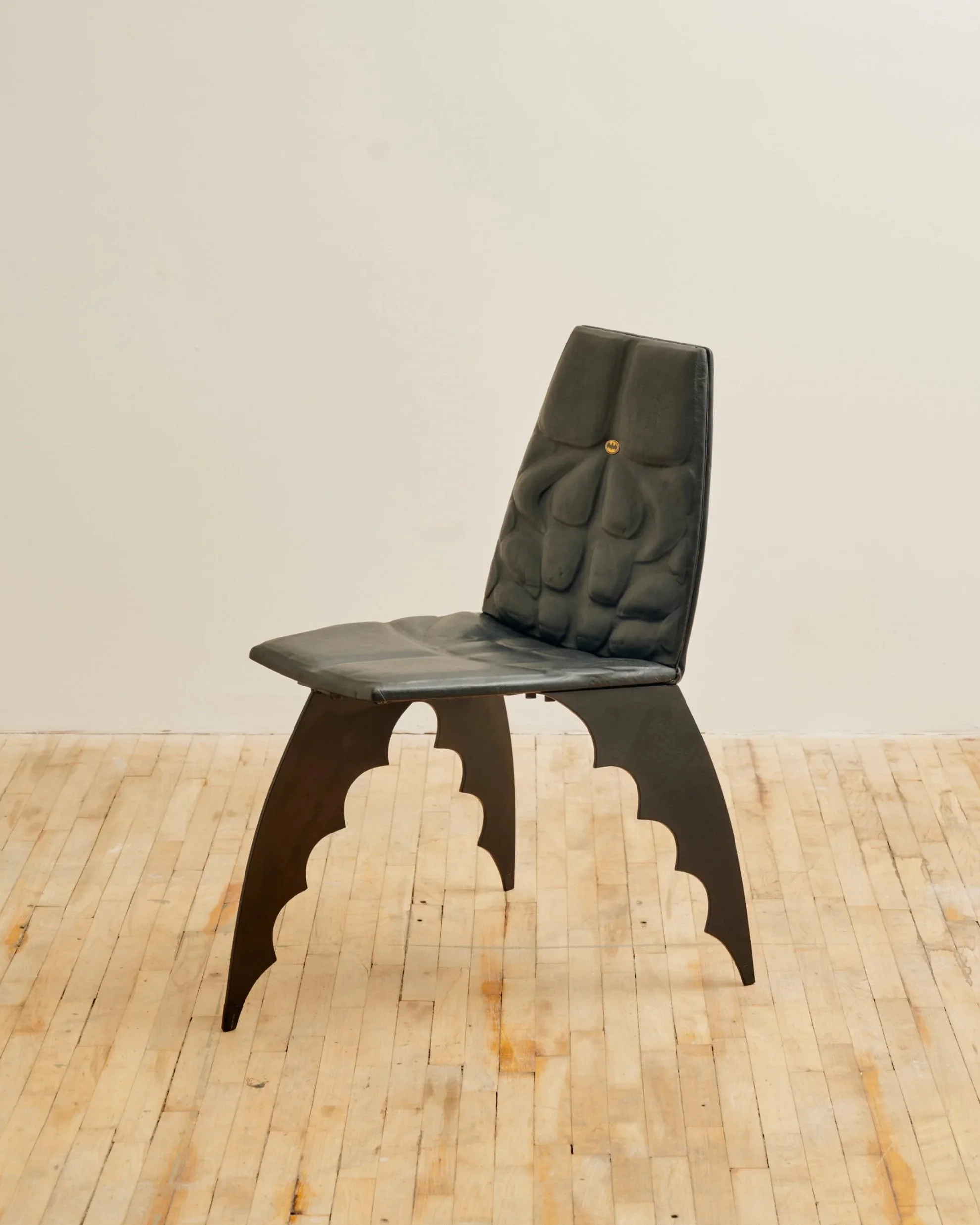 A black modern chair with a padded backrest and seat, featuring an unusual design on the legs, placed on a light wooden floor against a white wall.