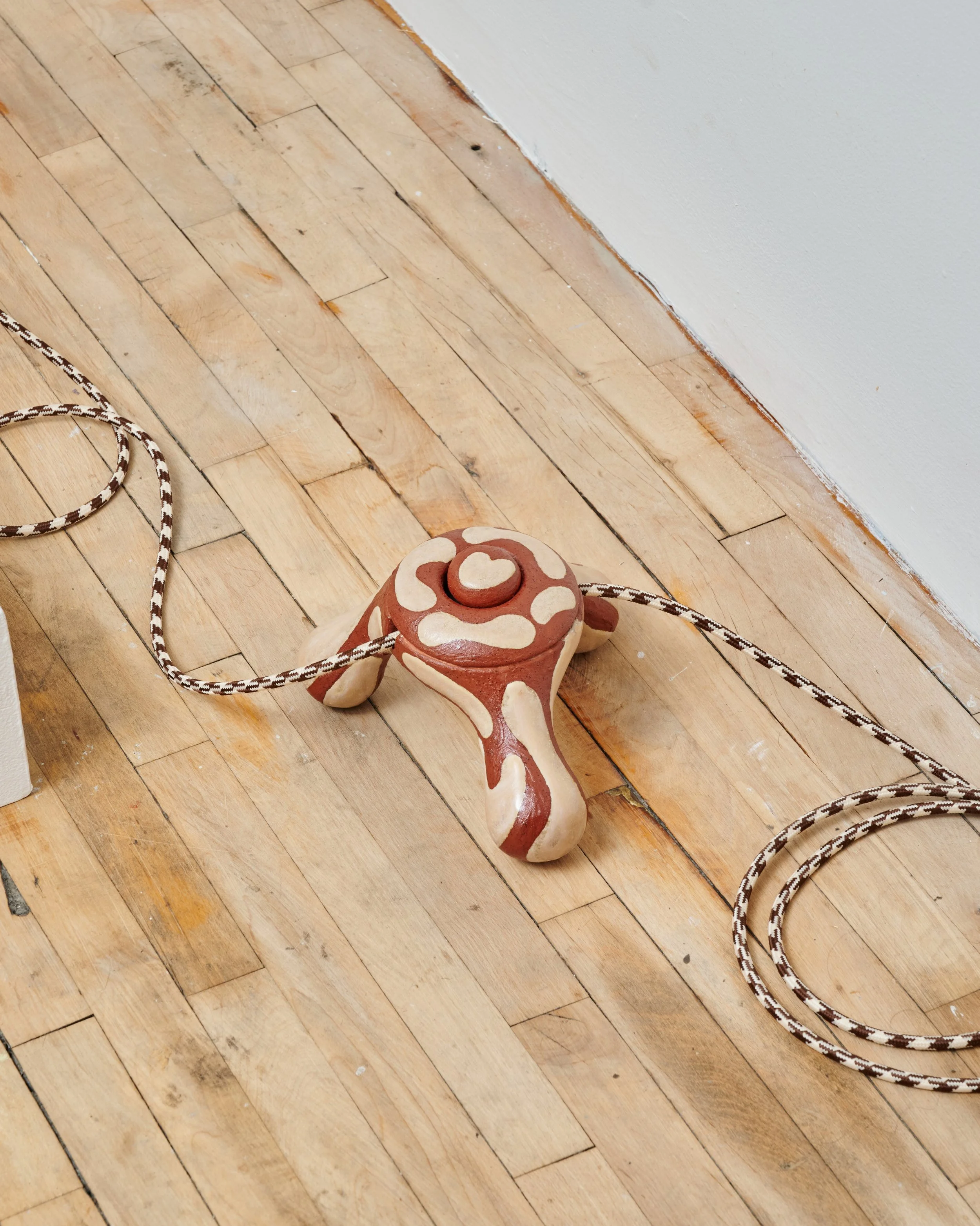 A terracotta-colored ceramic frog-shaped object with white and red decorative patterns, placed on a wooden floor with a white wall nearby. Joël Brodovsky-Adams.