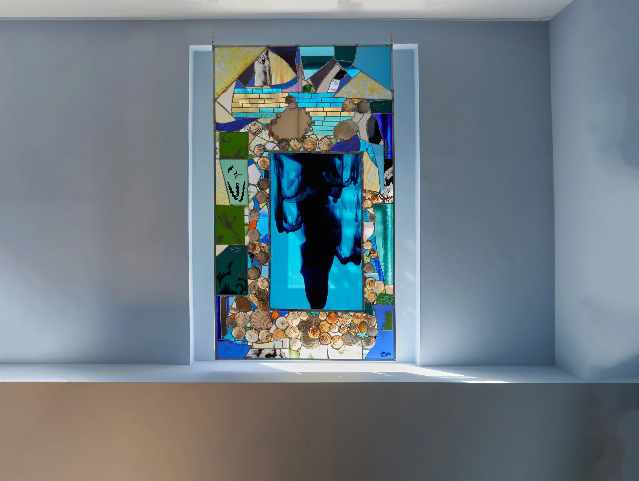A stained glass window with a central blue portrait of a woman, surrounded by various colorful geometric and abstract patterns, curated with beach and nature motifs, installed in a white wall.