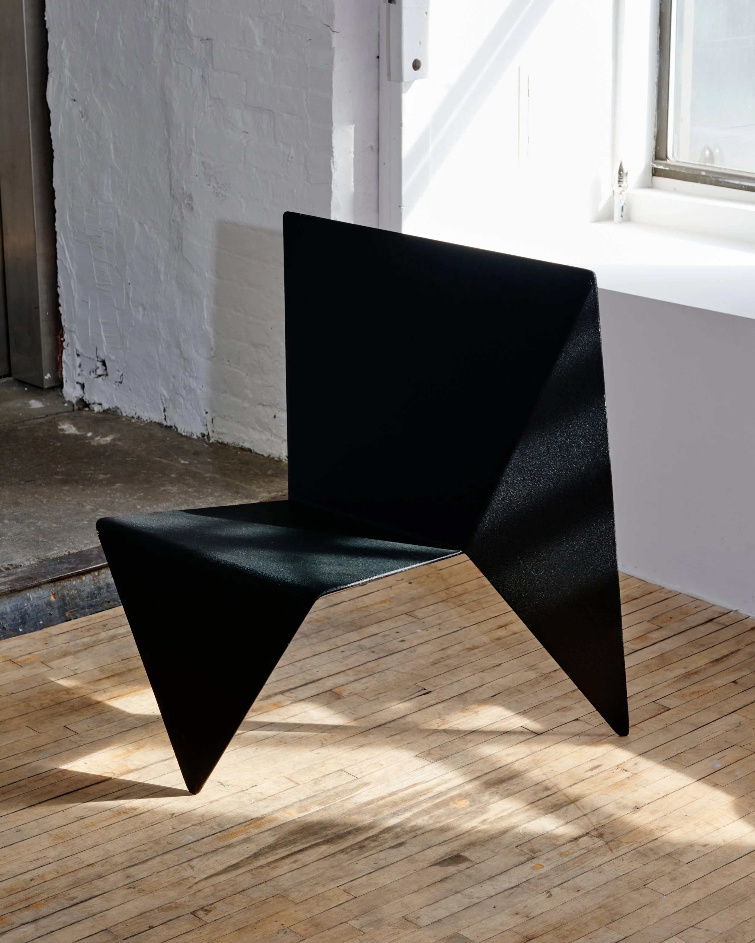 Black modern chair with angular design placed on wooden floor next to a white brick wall and near a window letting in sunlight.