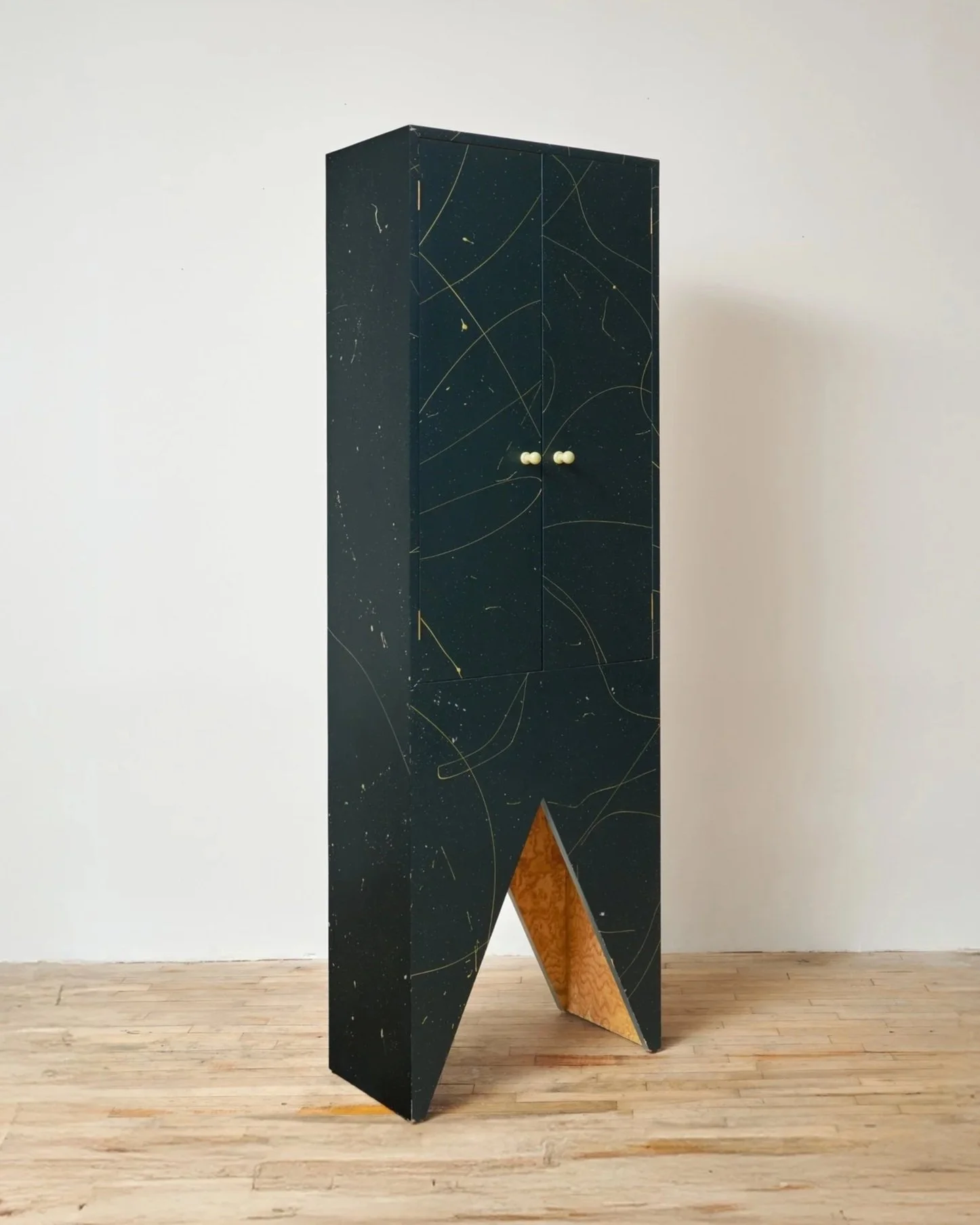 A tall, black cabinet with a wood interior at the bottom, decorated with gold lines and speckles, placed on a wooden floor against a plain white wall.