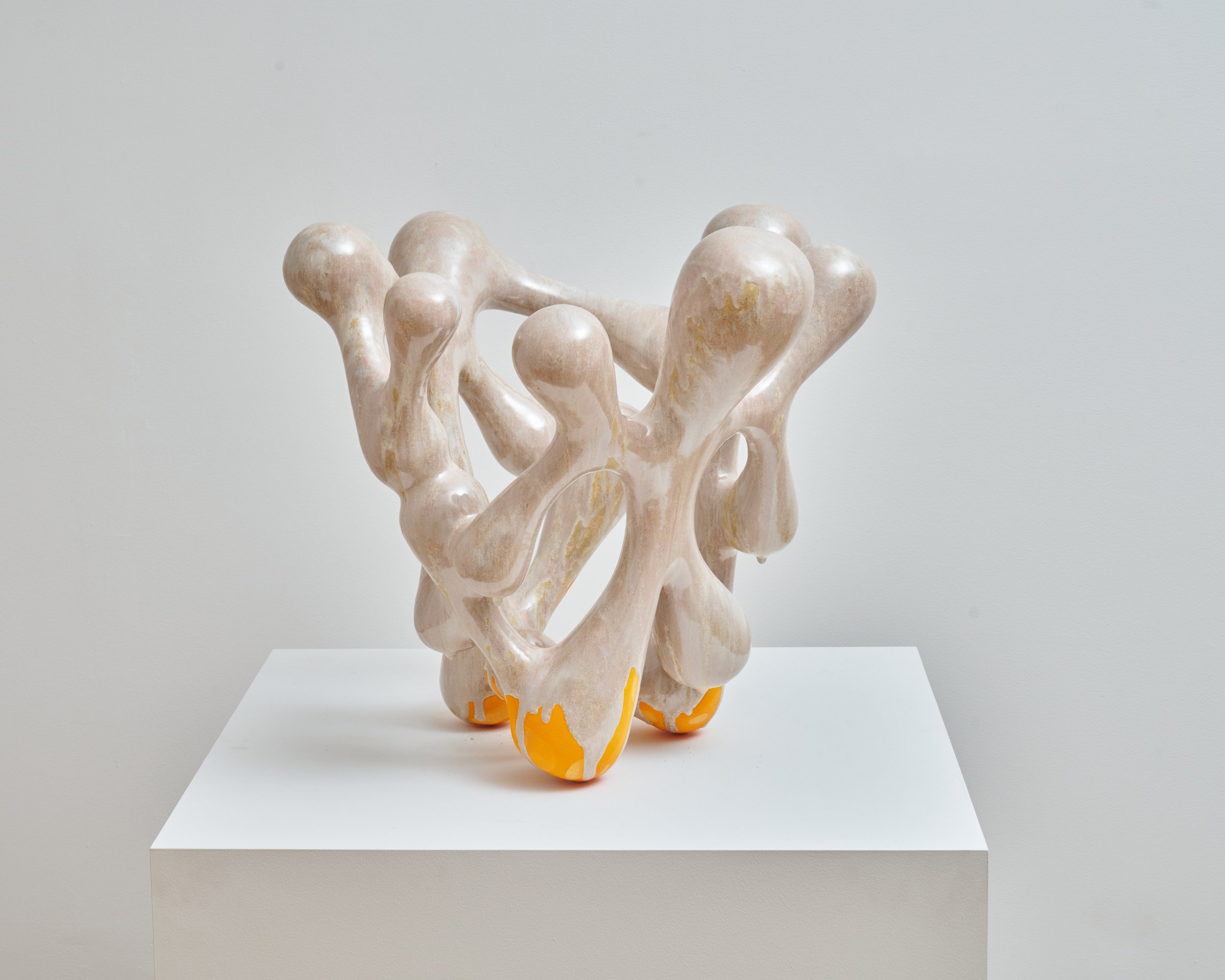 An abstract sculpture with interconnected, organic shapes in beige and orange, displayed on a white platform against a plain gray wall. Joël Brodovsky-Adams.