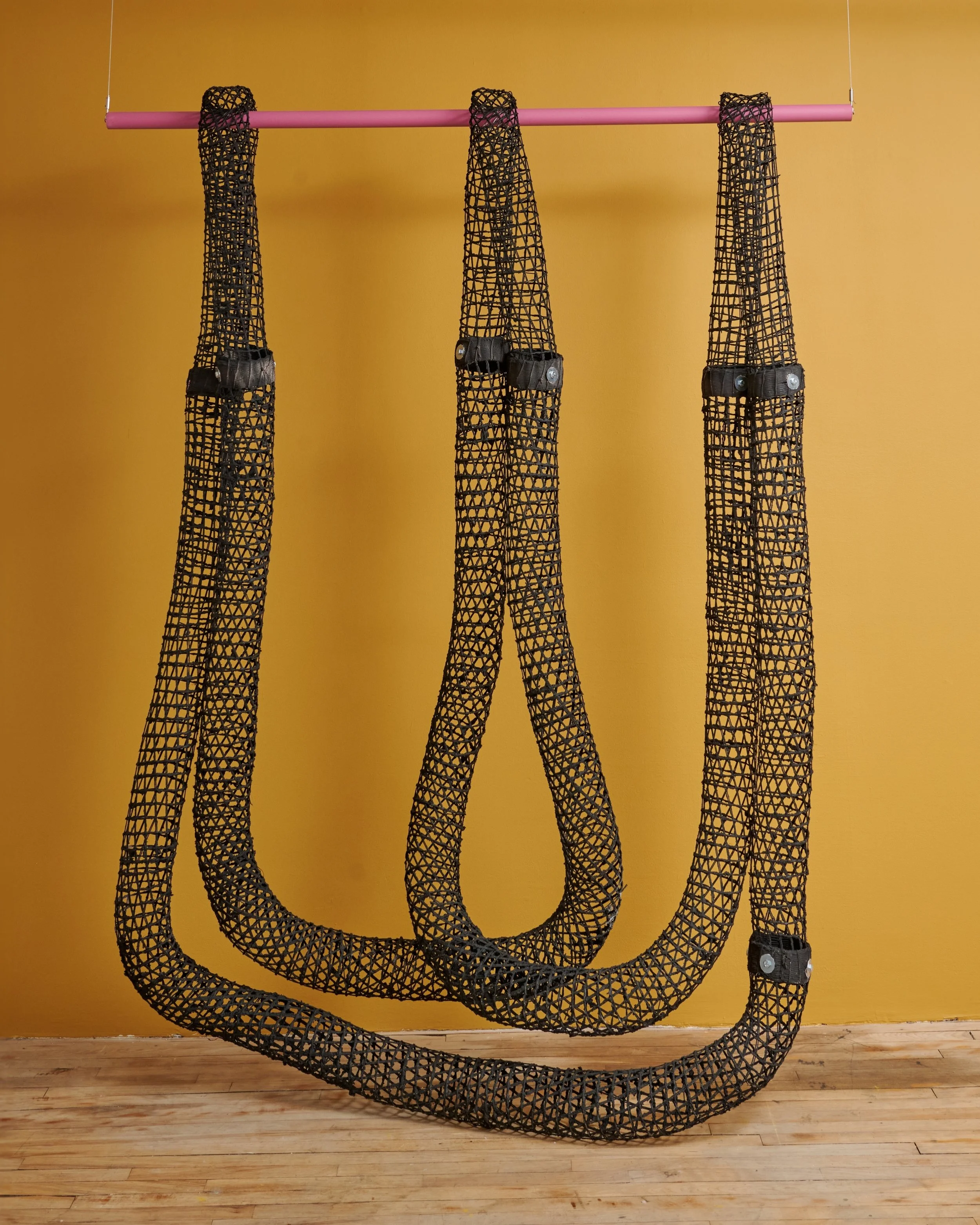 Large art installation resembling three intertwined black mesh tubes hanging from a pink rod, set against a yellow wall with a wooden floor.