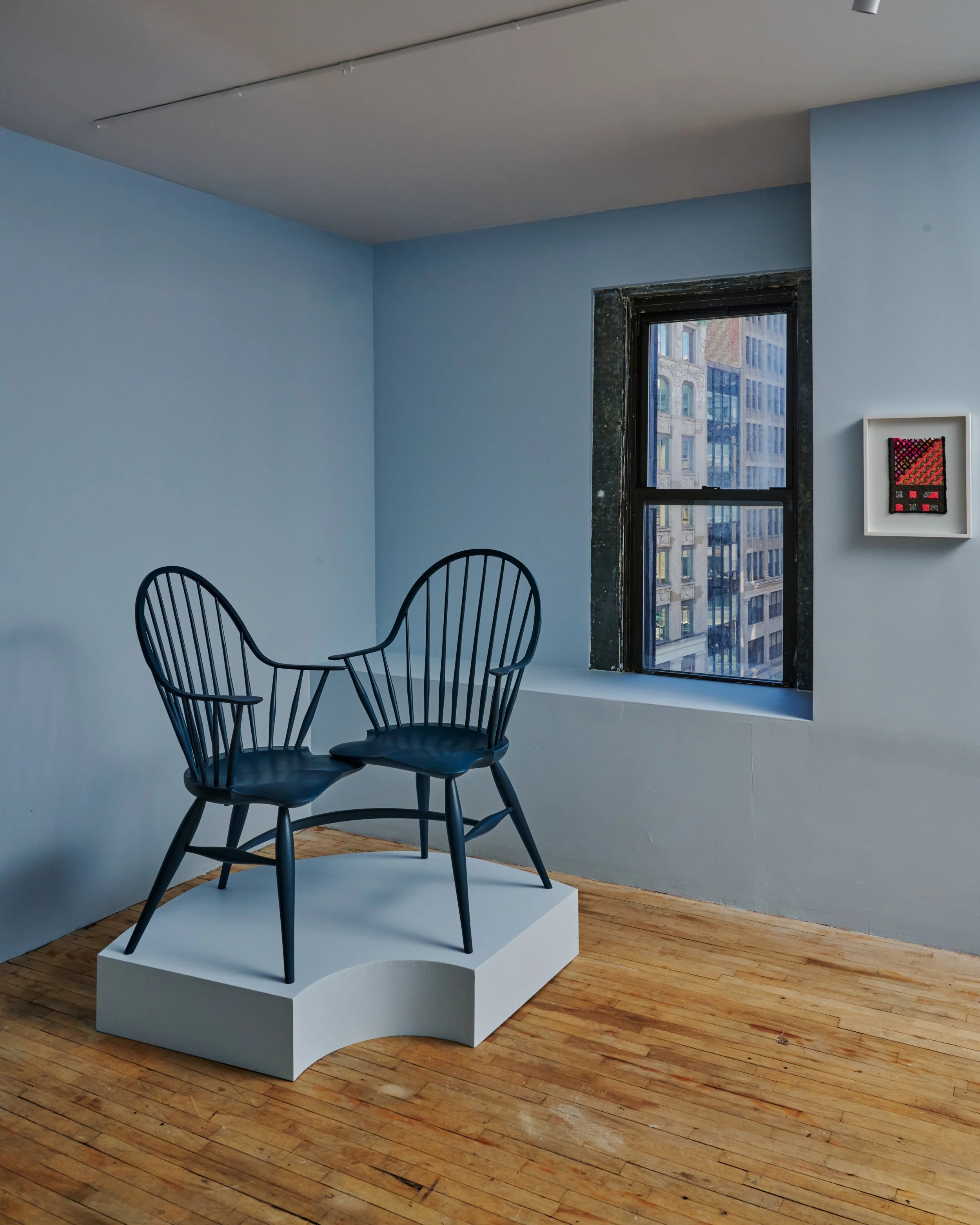 Two navy blue chairs with curved backs placed on a white platform in an art gallery with a wooden floor, a large window with black framing, and minimal wall decor.