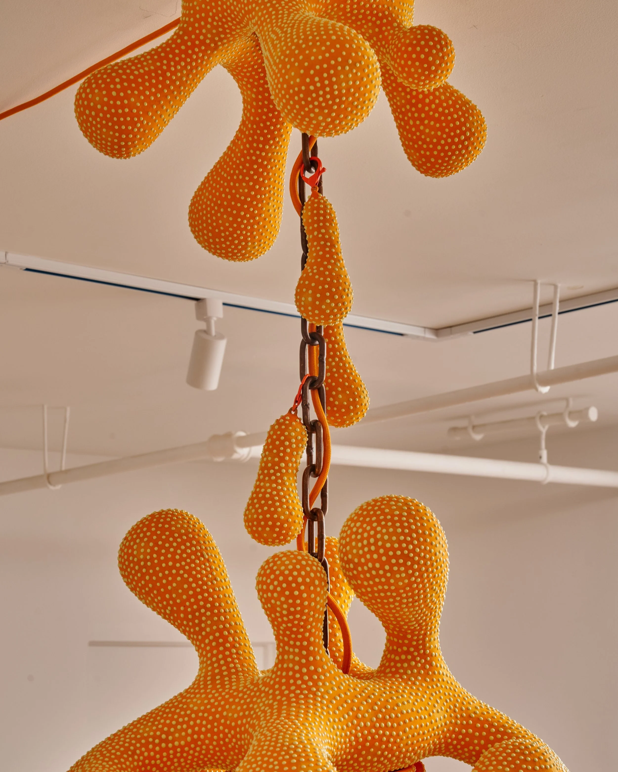 Close-up view of an orange and yellow dotted decorative sculpture resembling a stylized animal or abstract form, hanging from a chain. Joël Brodovsky-Adams.
