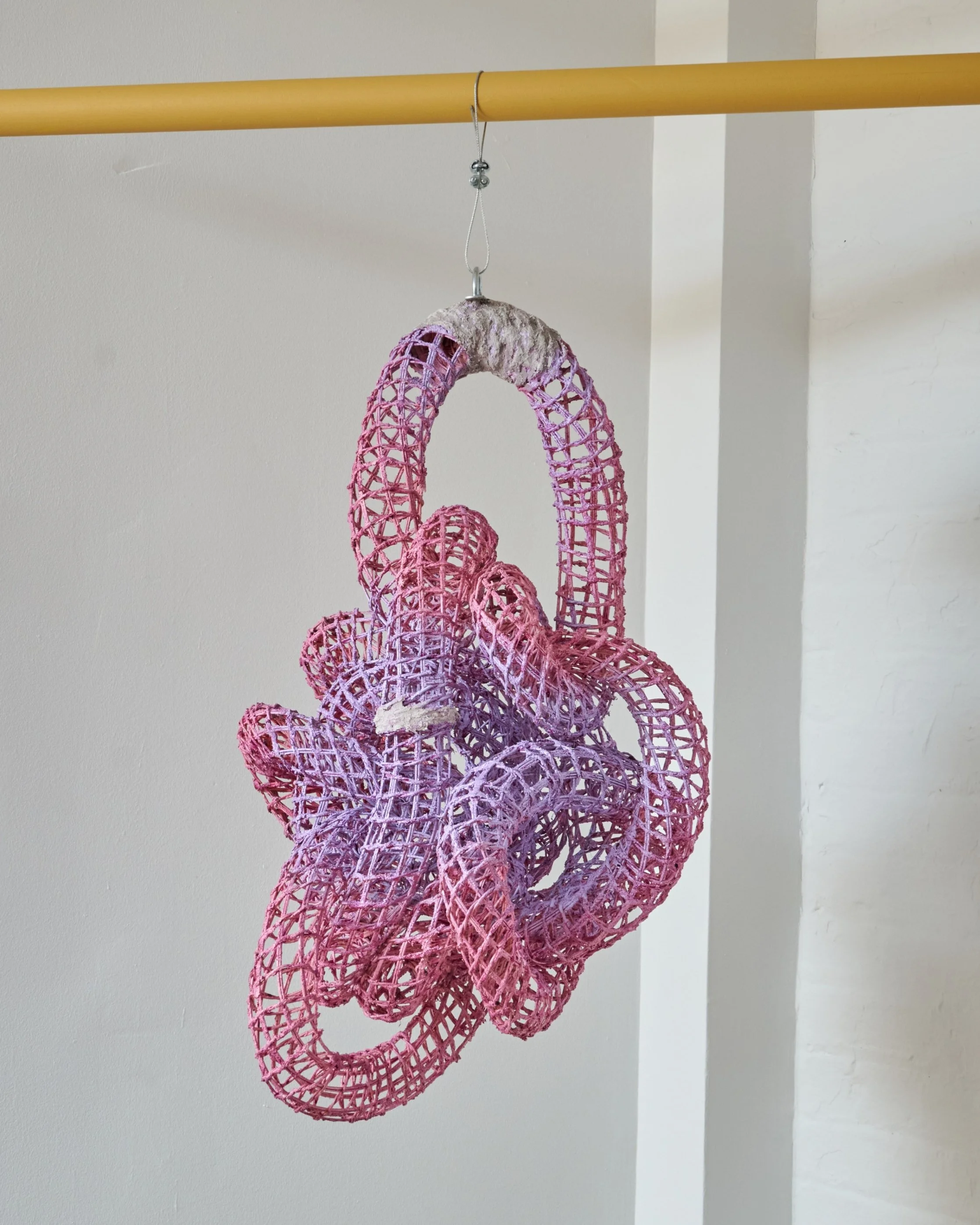 A colorful, abstract chandelier made of intertwined pink and purple woven tubes hanging from a ceiling with a yellow pole.
