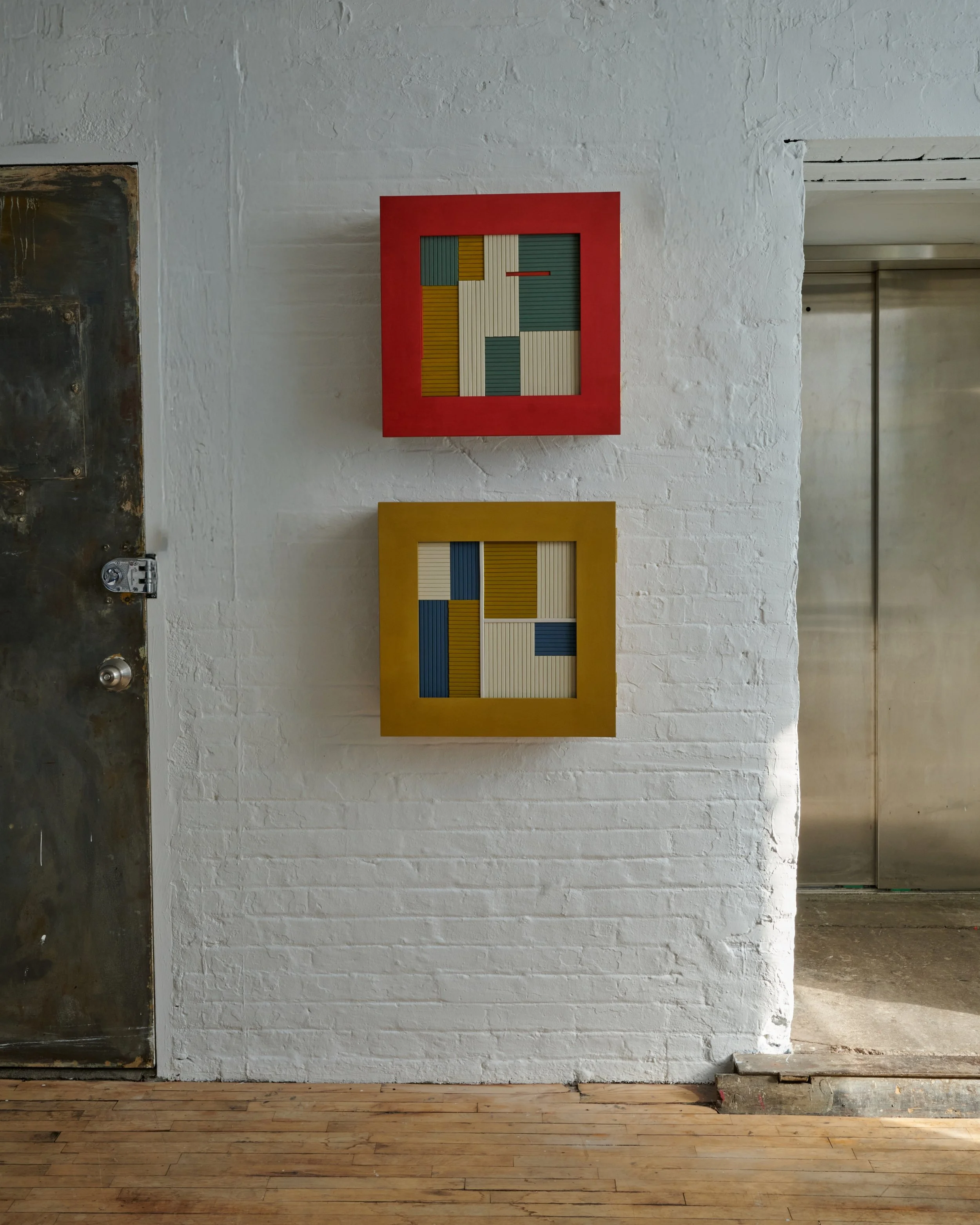 Two colorful abstract art pieces with geometric patterns hanging on a white brick wall, one with a red frame and the other with a yellow frame.