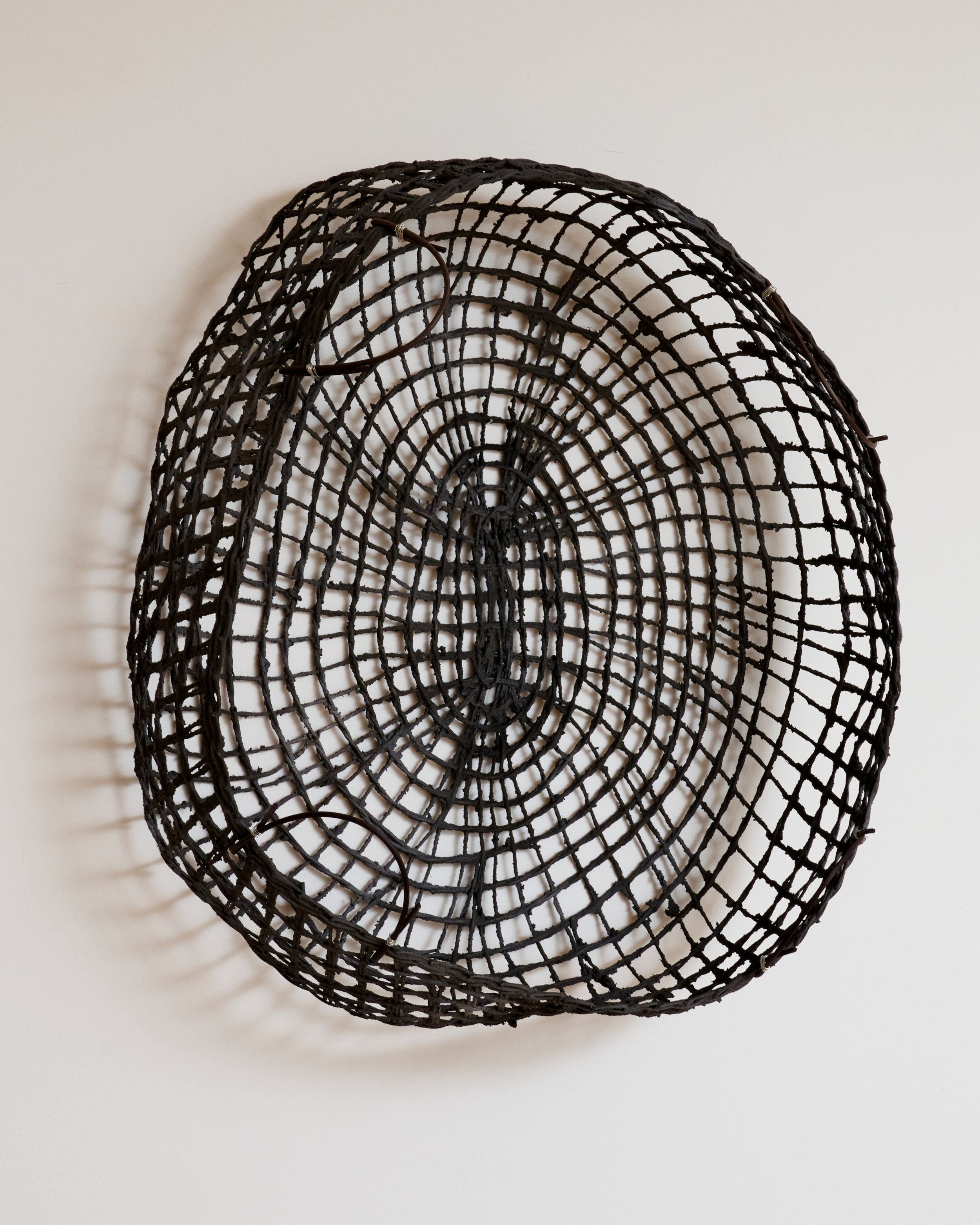 A black, woven, circular basket with a spiral pattern hangs on a white wall.